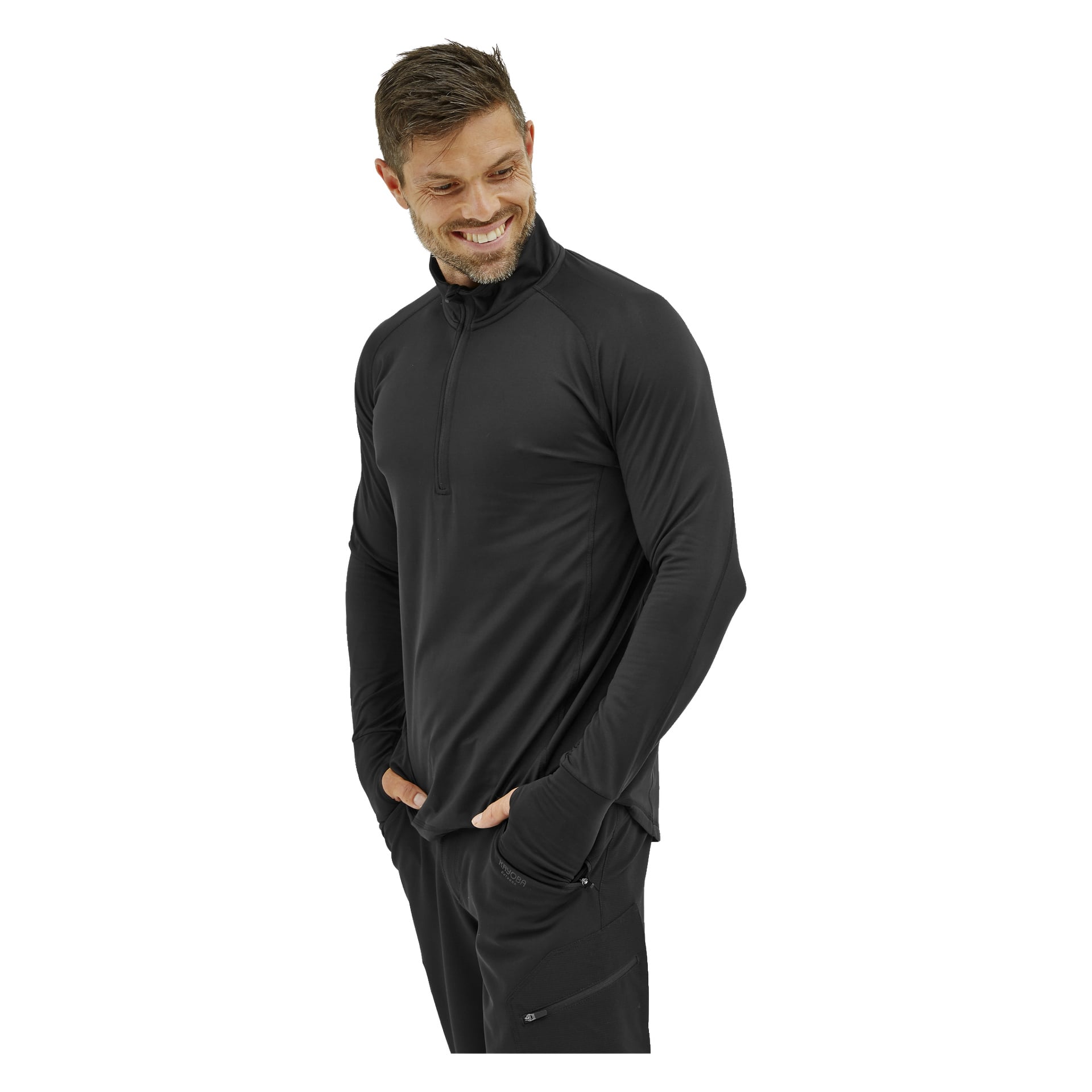 Ein Mann mit kurzen braunen Haaren und Bart trägt das Kayoba Men's Functional Top London in Schwarz, kombiniert mit schwarzer Hose. Er steht mit den Händen in den Taschen, schaut nach links und lächelt.