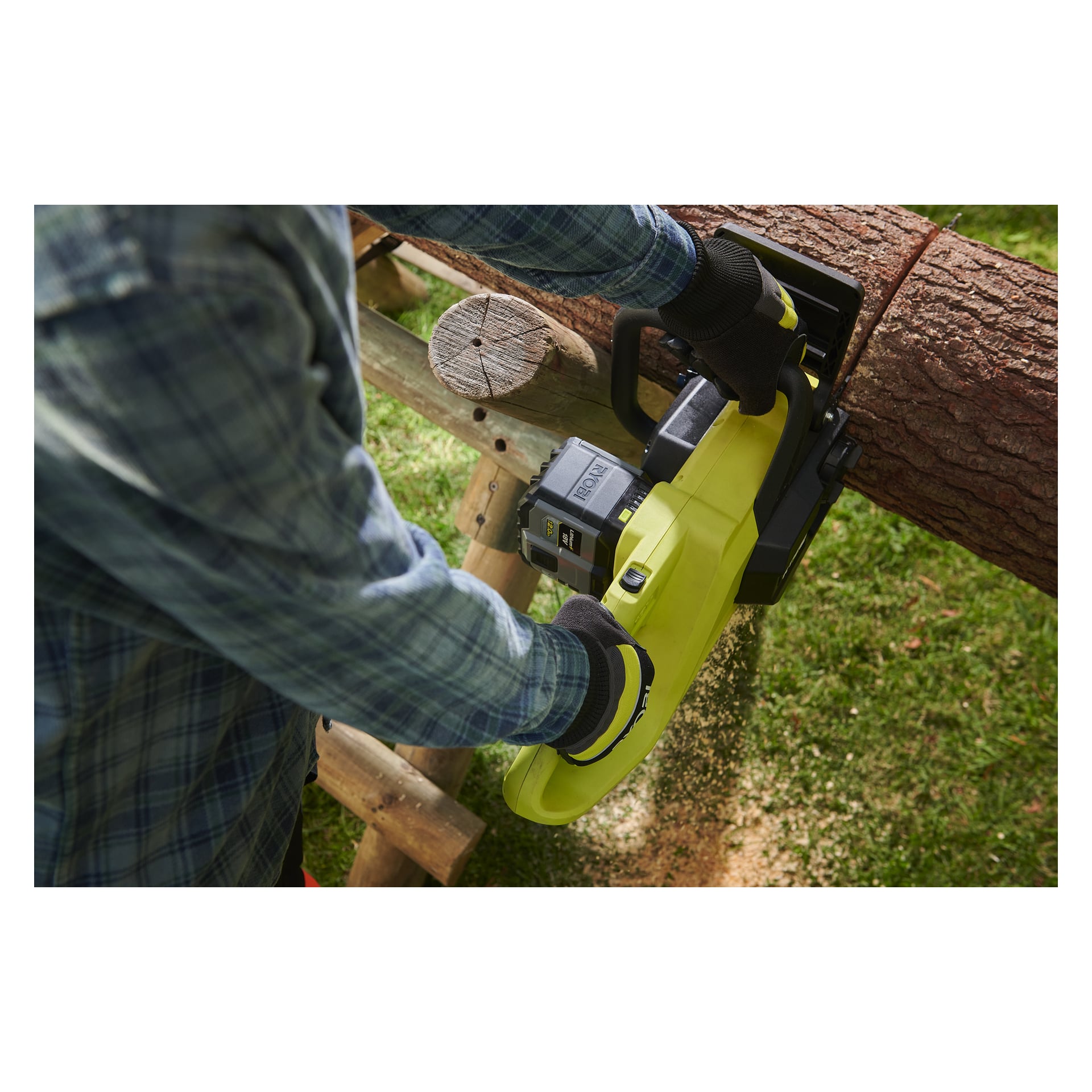 En person med hansker og rutete skjorte bruker en gul Ryobi One+ Battery High Energy RB18120T elektrisk motorsag for å kutte en stokk på en tresag utendørs, med sagflis spredt nær kuttet.