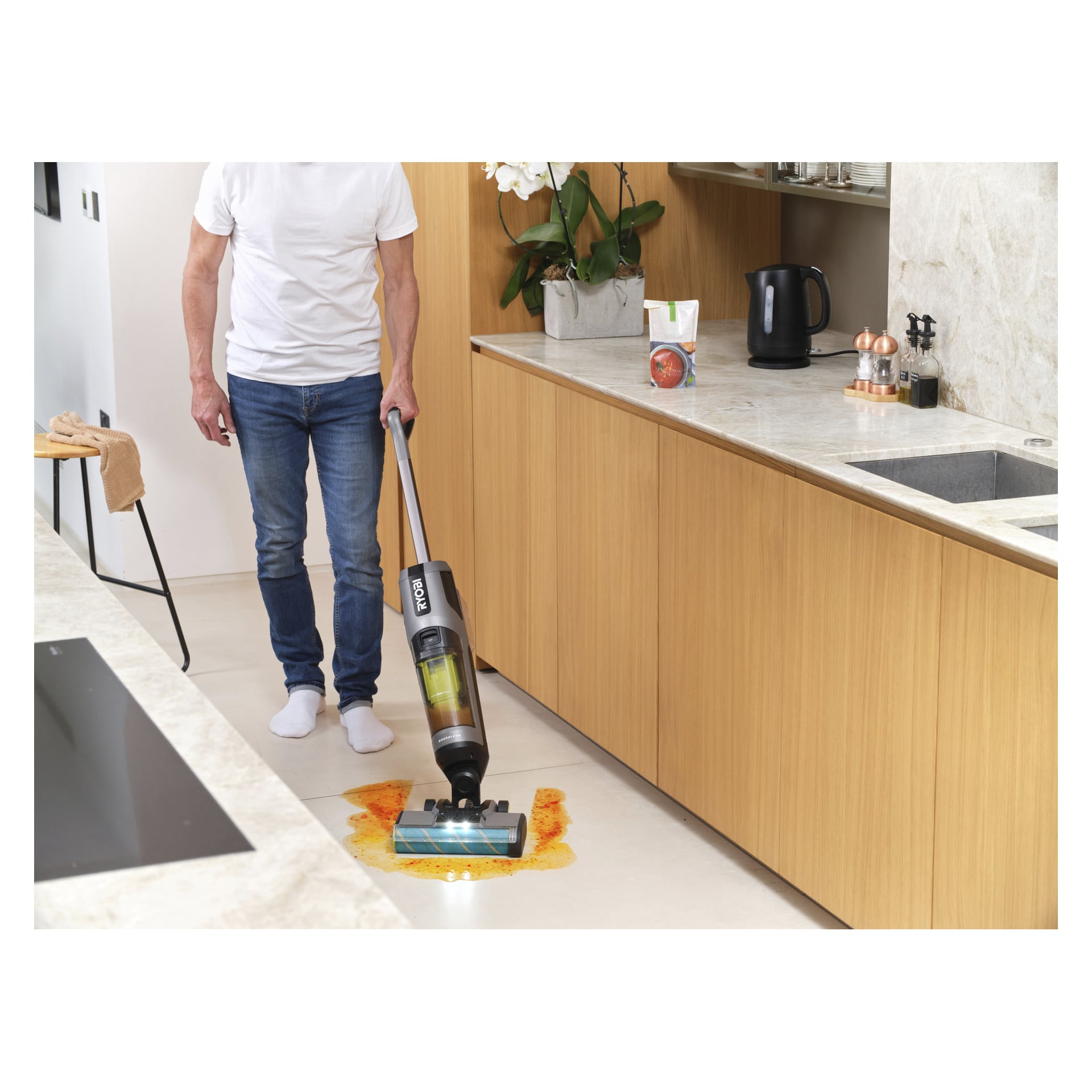 Eine Person, die ein weißes Hemd und eine weiße Jeans trägt, benutzt den Ryobi One+ 3-in-1 Upright Staubsauger mit Mopp-Funktion (RHFC18BL-0), um eine große orangefarbene Flüssigkeit auf dem Küchenboden zu beseitigen, umgeben von Holzschränken und mit einer Marmorarbeitsplatte bedeckt.