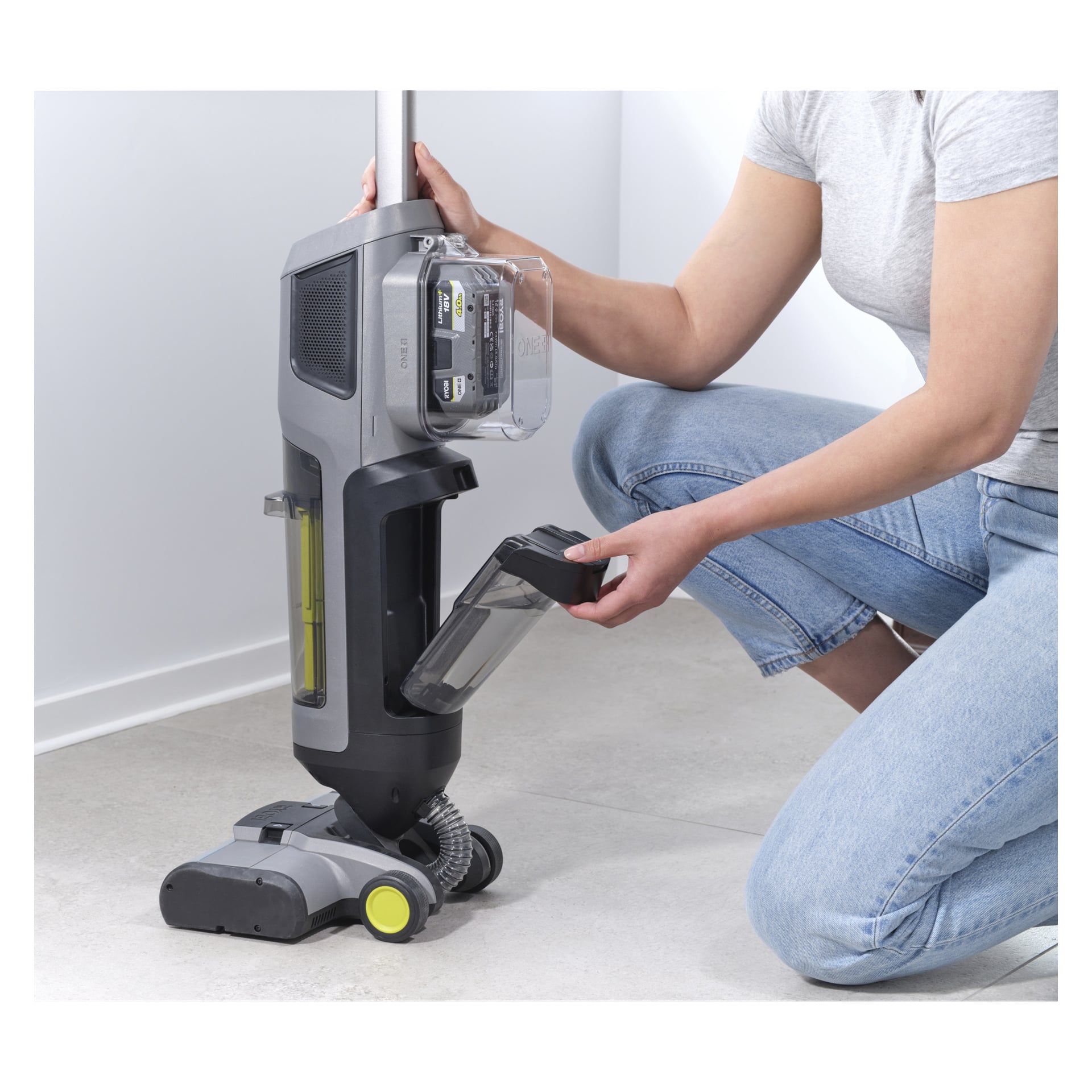 Eine Person in grauem Hemd und blauen Jeans kniet auf einem gefliesten Boden und nimmt den Mülleimer aus dem aufrechten Ryobi One+ 3-in-1 Staubsauger mit Wischfunktion, Modell RHFC18BL-0.