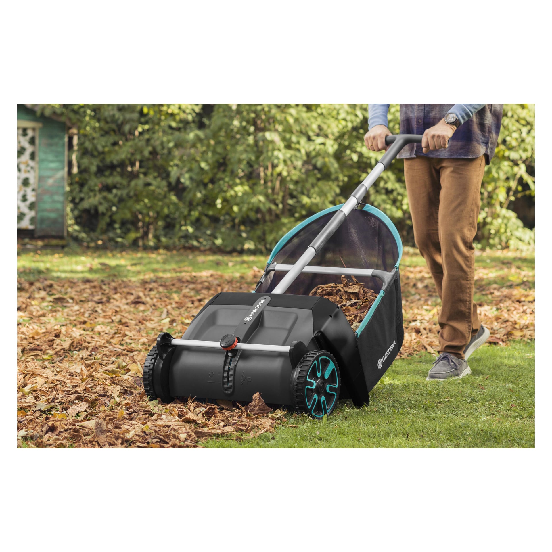 Jemand bedient den Gardena Leaf Collector mit seinen schwarzen und türkisfarbenen Akzenten, um Blätter aufzusammeln, die über einen grasbewachsenen Garten verstreut sind, gekleidet in braune Hosen und graue Schuhe.