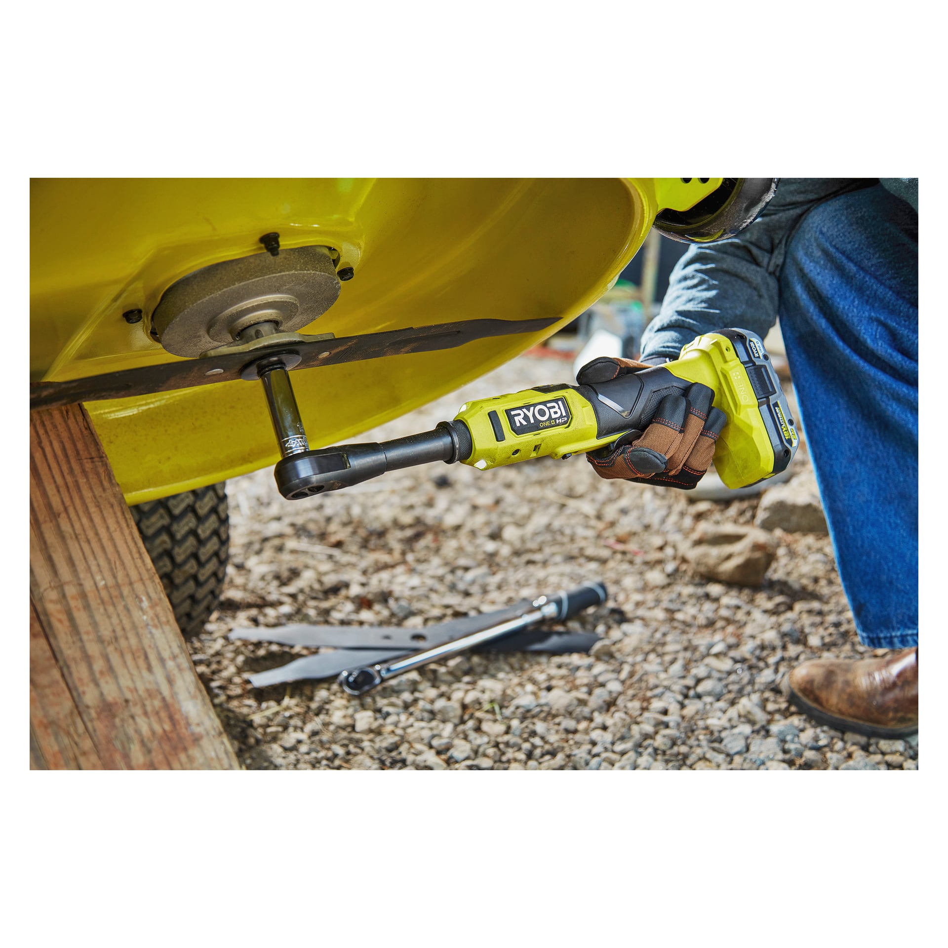 Ktoś obsługuje na baterii Ryobi One+ HP grzechotkę RRW1838X-0 na śrubie przymocowanej do żółtej metalowej powierzchni na zewnątrz, stojąc na żwirze z innymi porozrzucanymi narzędziami w pobliżu.