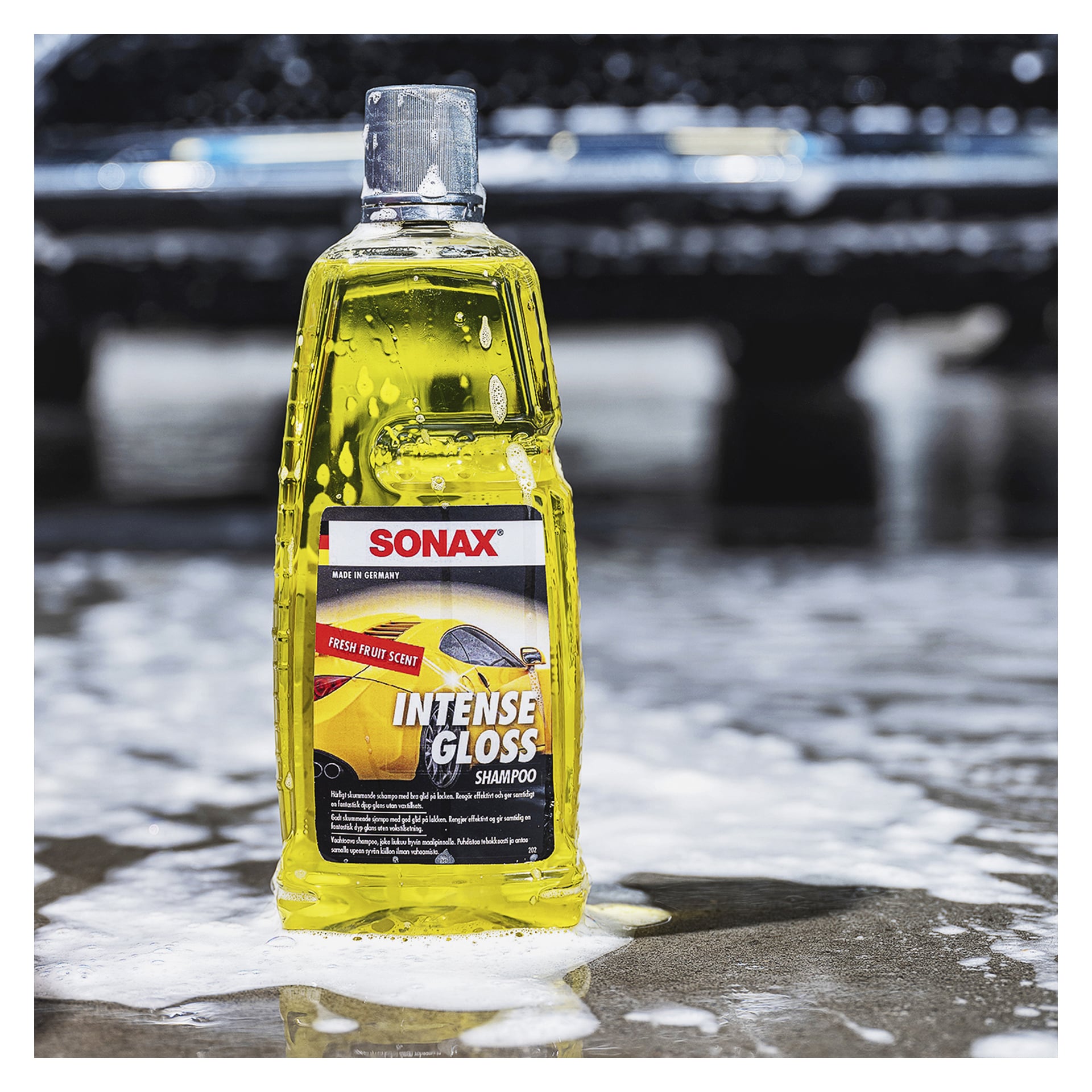 En gul flaska märkt "Car shampoo Intense gloss" från Sonax står på våt asfalt med tvålskum runt omkring, och en suddig svart bil i bakgrunden.