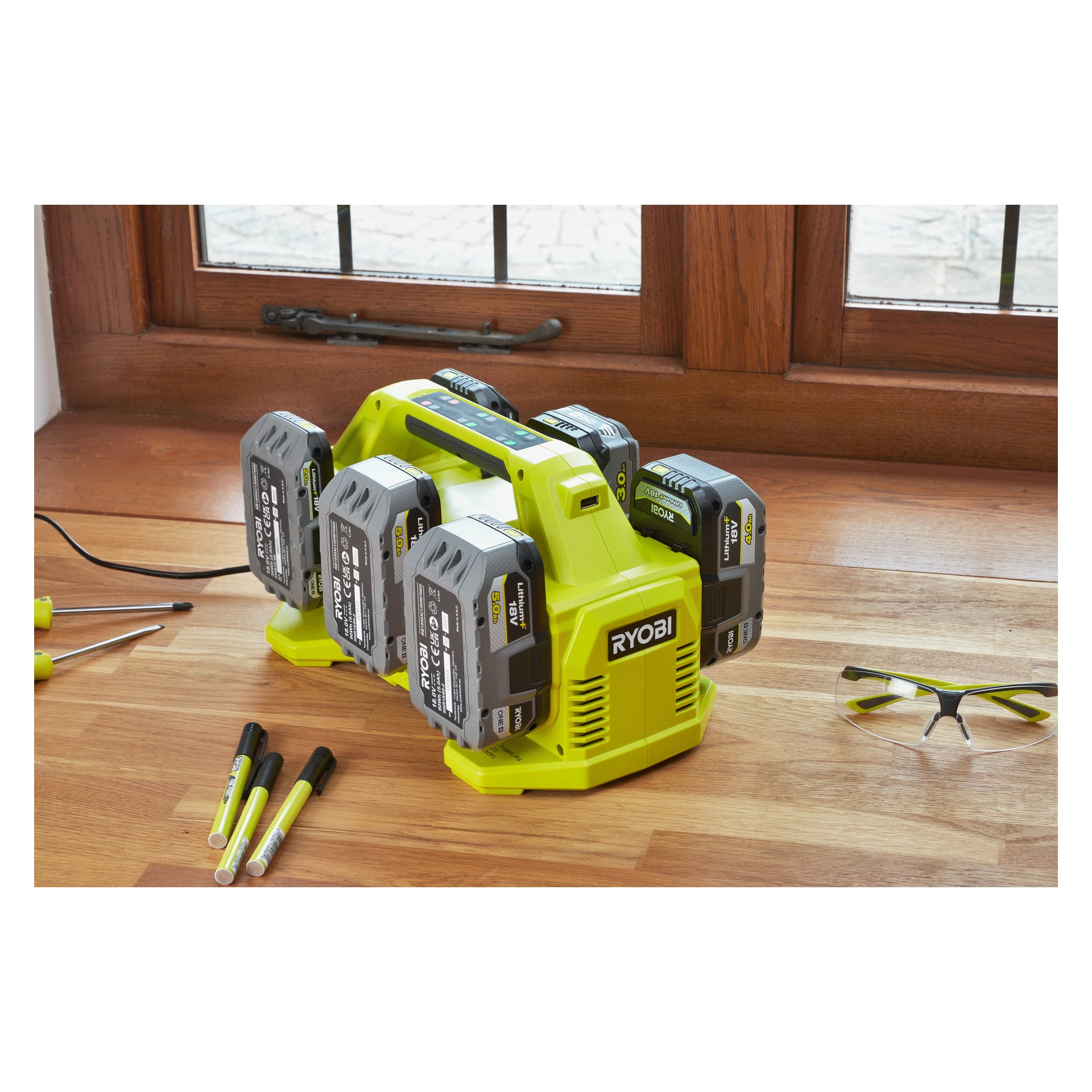 Limenvihreä Ryobi One+ Multi-laturi (malli RC18640) lataa neljää litium+ 18V akkua. Laturi lepää puupinnalla ikkunan vieressä, ympärillään suojalasit, kolme merkkiä ja kaksi ruuvimeisseliä.