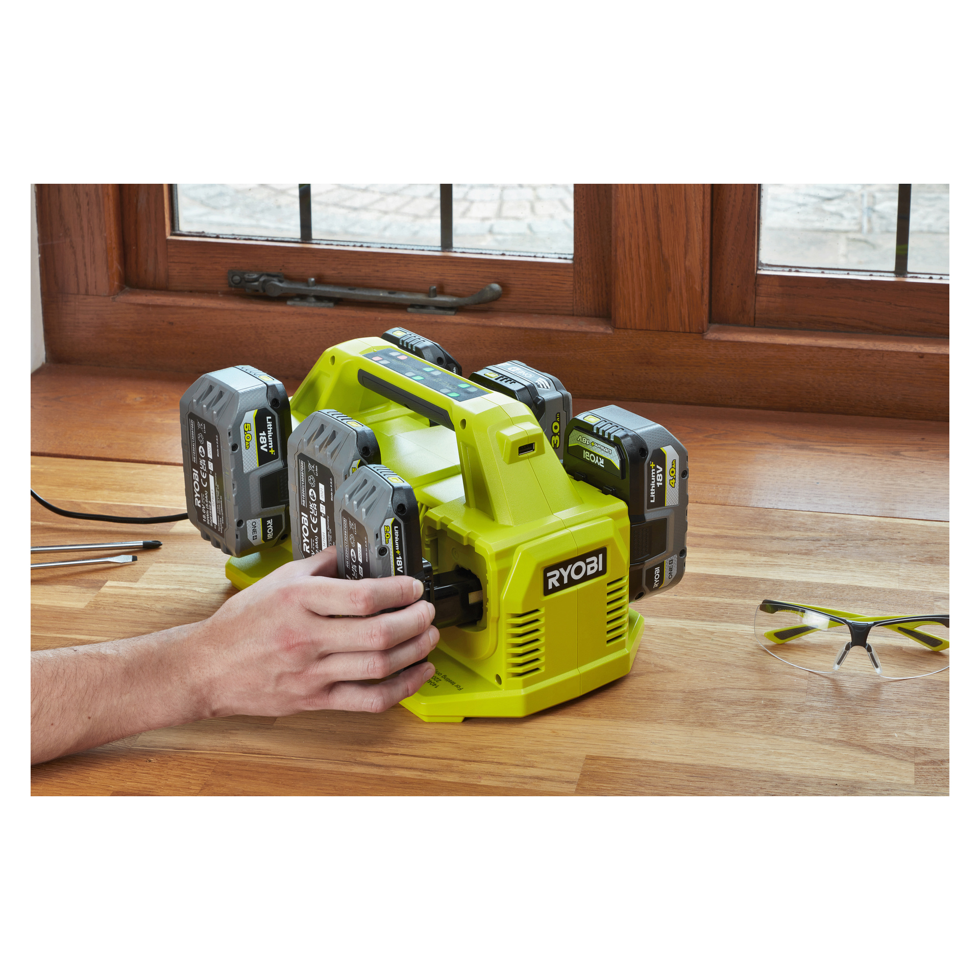 Joku liu'uttaa Ryobi One+ -akkua Ryobi One+ Multi -laturin RC18640 sisään, joka on suunniteltu kuudelle akulle ja lepää puupinnalla. Yksikössä ladataan jo kolme muuta akkua, ja lähellä on turvalasit sekä erilaisia pieniä työkaluja.