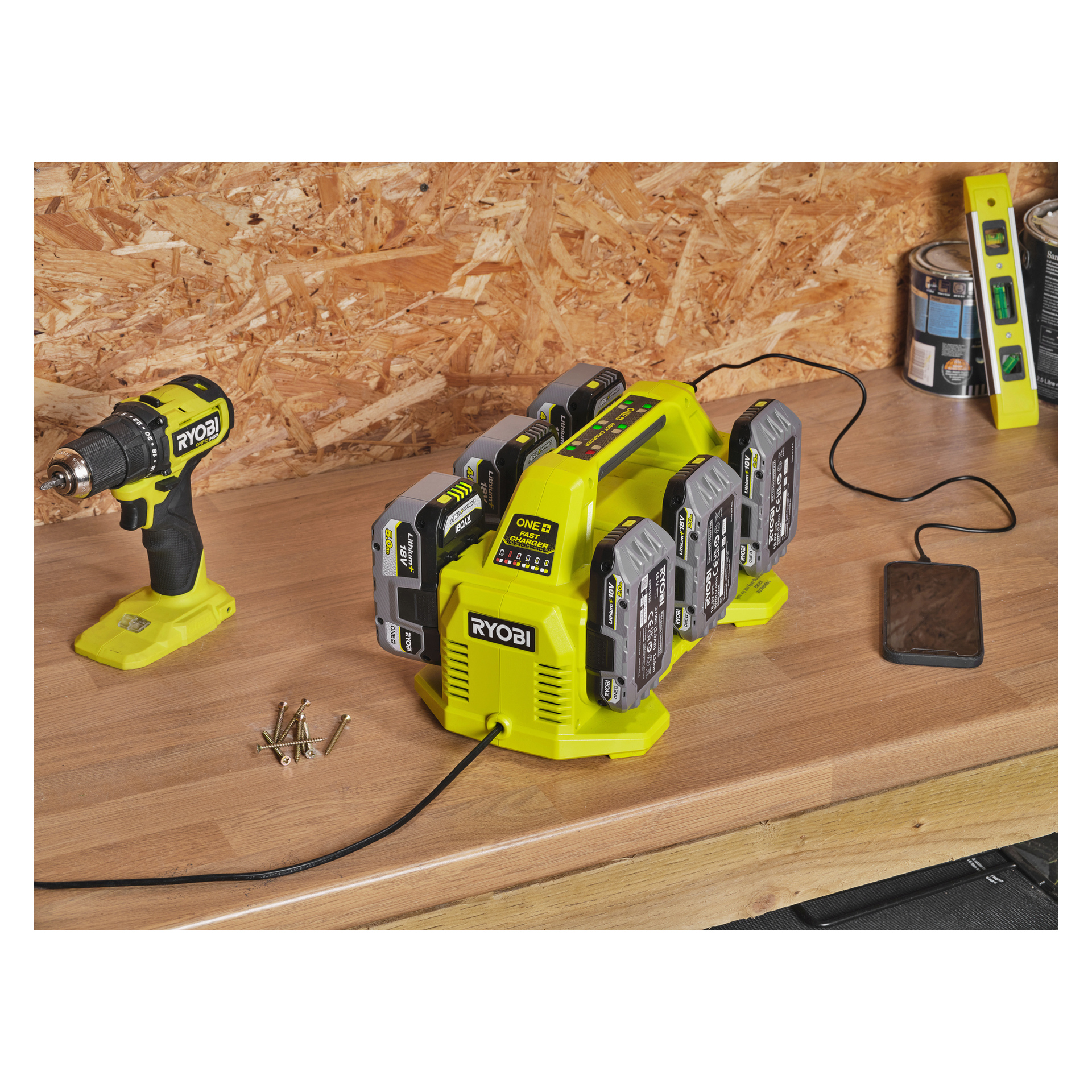 Ryobi One+ Multi -laturin RC18640, joka on suunniteltu kuudelle akulle, lepää puisella työpöydällä, jossa on useita akkuja latauksessa. Vasemmalla puolella näkyy johdoton pora ja ruuvit; Oikealla puolella on puhelin ja henkitaso. Taustana on suunnattu levyseinä.