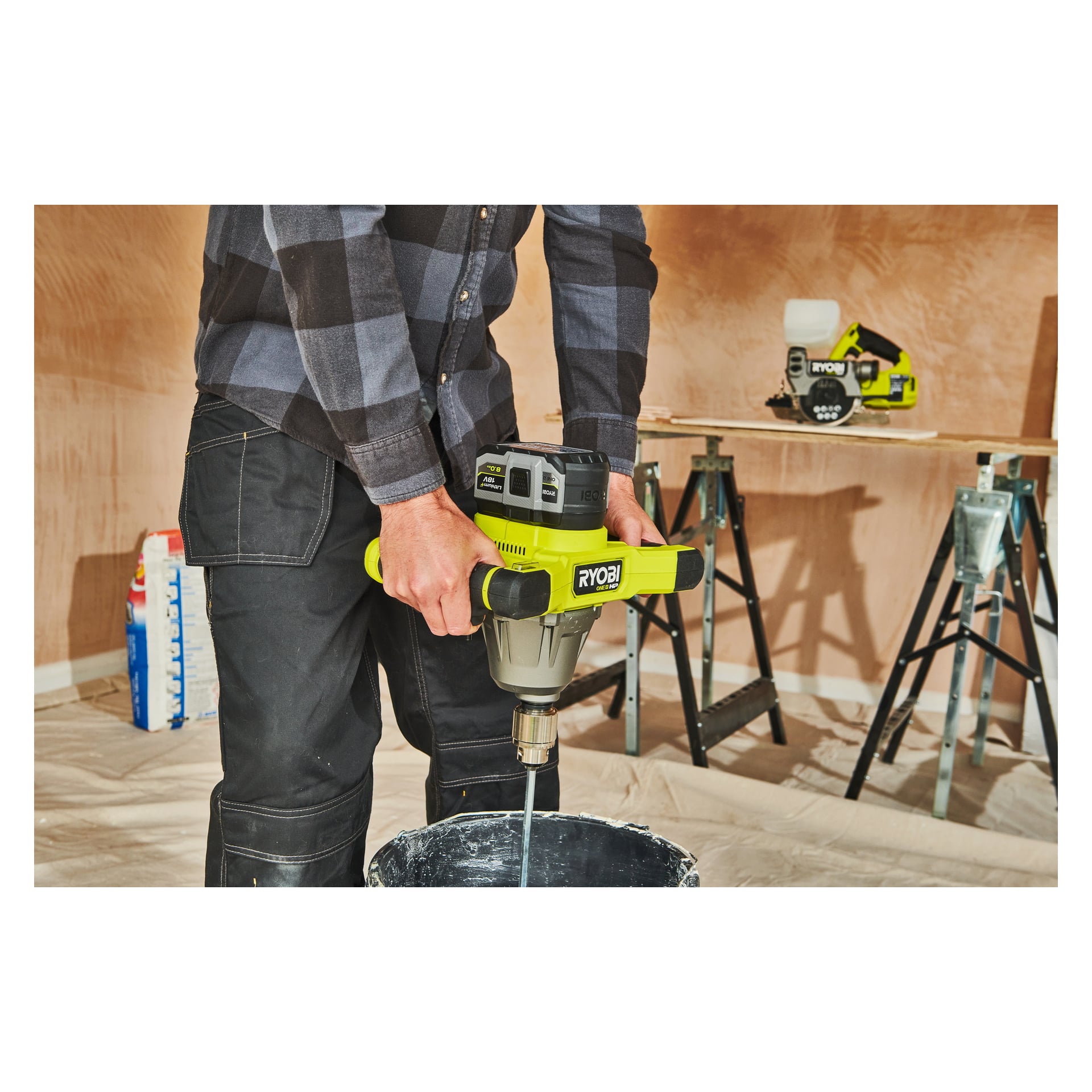 En person som bär en rutig skjorta använder Ryobi One+ Battery High Energy RB1880T med en kraftblandare för att blanda material i en hink. Ett annat Ryobi-verktyg står på en arbetsbänk i närheten på en renoveringsplats.
