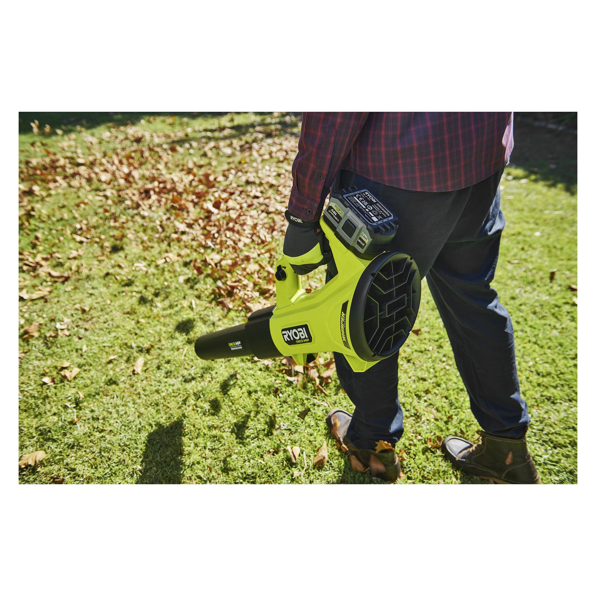 En person som håller i en gul "Ryobi One+ Battery High Energy RB1880T"-lövblåsare står på ett gräsområde täckt av fallna löv, klädd i bruna skor, mörka byxor och en rutig skjorta.