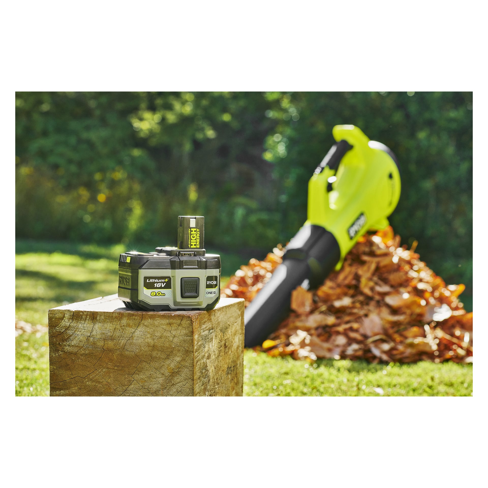 En Ryobi One+ Battery High Energy RB1880T och laddare står på en trädstubbe, medan en Ryobi-lövblåsare står på höstlöv i den soliga trädgården bortom.