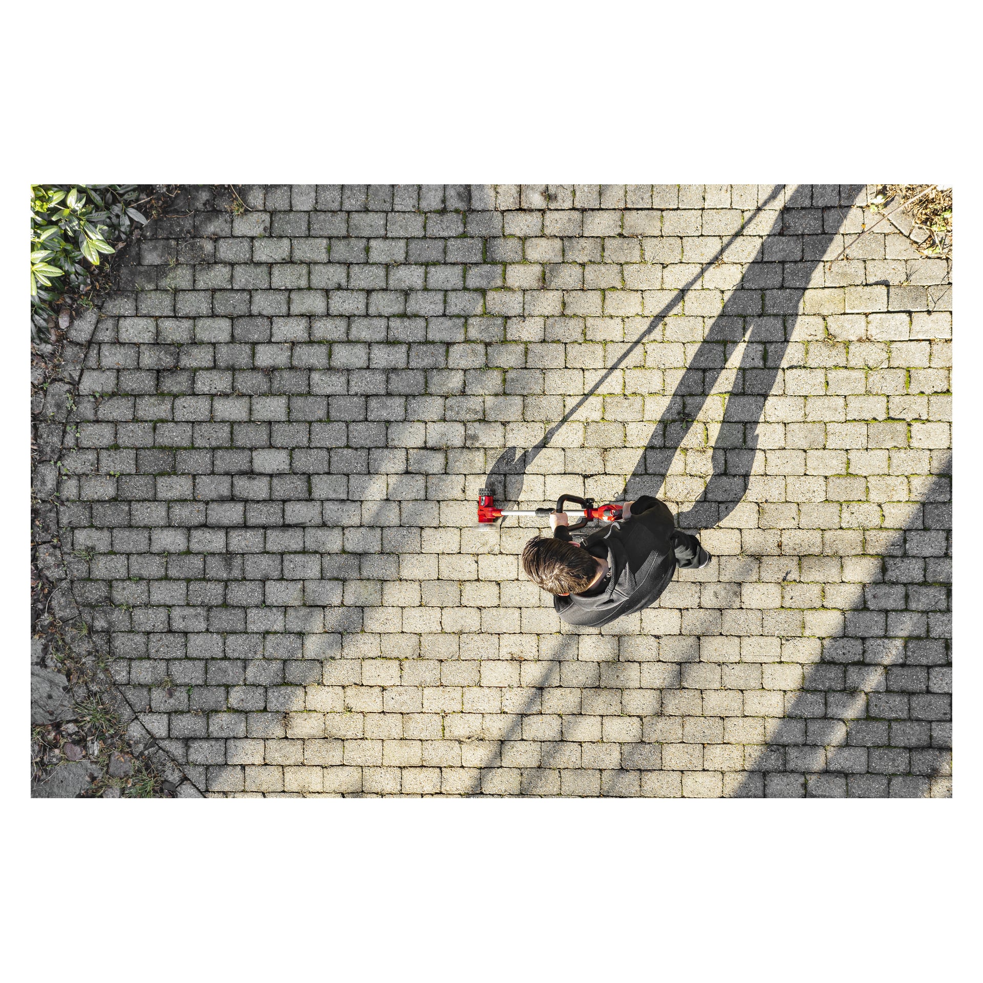 Z góry osoba w ciemnym ubraniu obsługuje Meec Tools Multiseries Paving Scraper PW-18V HOME 300 na prostokątnej ceglanej nawierzchni. Długie cienie opadają na teren, a na jednej stronie rosną rośliny.