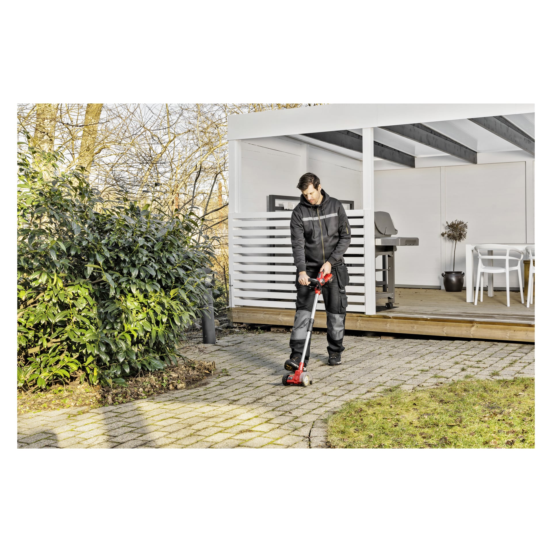 Ubrany w ciemną odzież roboczą, osoba obsługuje czerwony Meec Tools Multiseries Paving Scraper PW-18V HOME 300, aby posprzątać lub objąć kamienną ścieżkę obok nowoczesnego patio z białymi meblami ogrodowymi i bujną zielenią otaczającą okolicę.