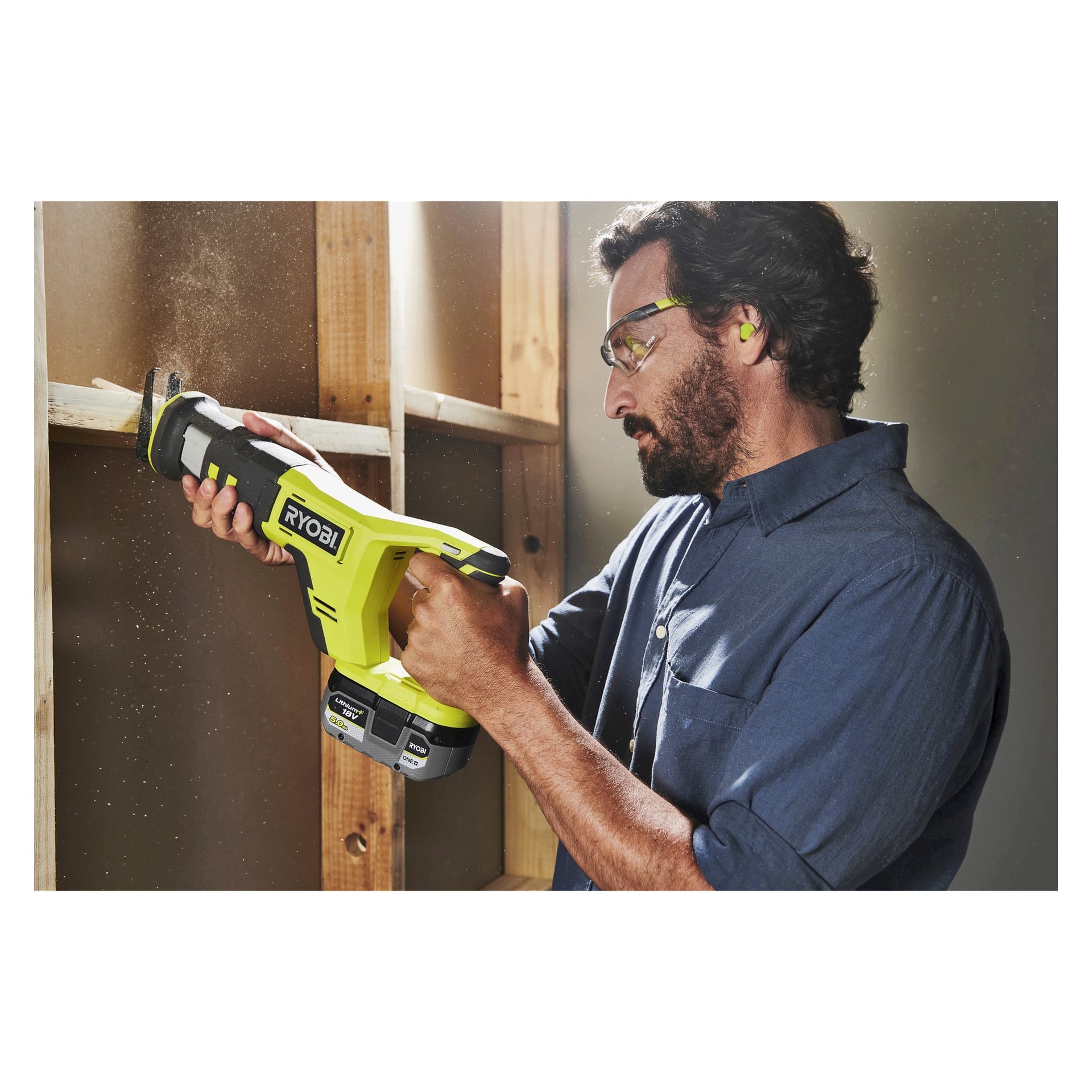 Ein Mann, geschützt durch Schutzbrillen und Ohrstöpsel, bedient eine gelbe Ryobi One+ batteriebetriebene Hubkolbensäge (Modell RRS18-125G), um einen Holzbalken in einer teilweise gebauten Wand zu durchschneiden, wobei um ihn herum Sägemehl herumflog.