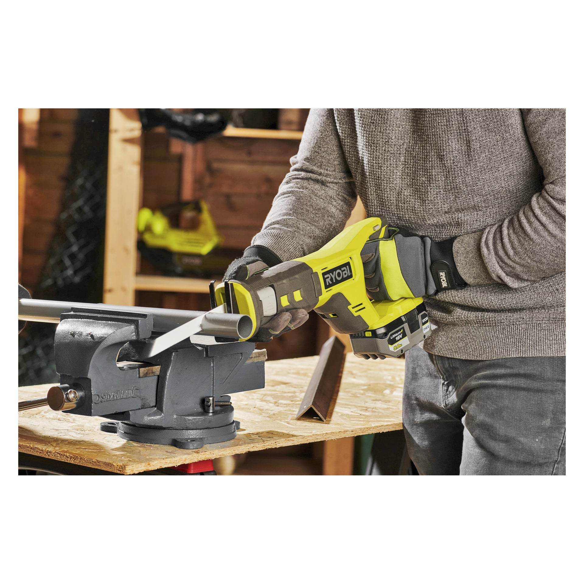 Handschuhige Hände bedienen eine batteriebetriebene Ryobi One+ Hubkolbensäge RRS18-125G, die durch ein Metallrohr schneidet, das von einem Schraubstock auf einer hölzernen Werkbank in einer Werkstatt gehalten wird.