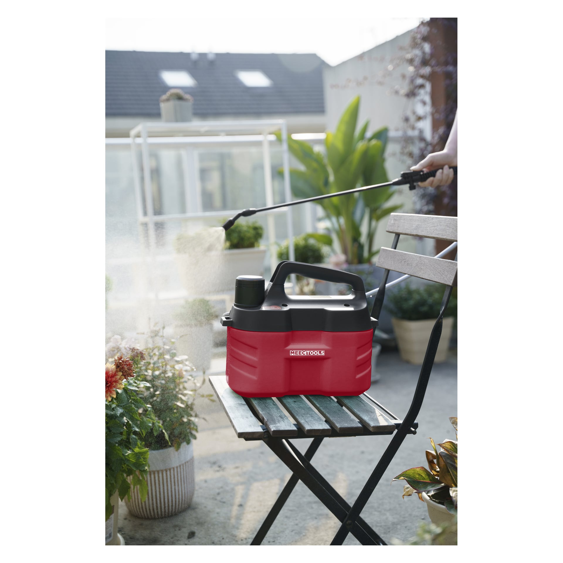 Eine Person sprüht in einem Garten, wo eine rot-schwarze, batteriebetriebene Meec Tools Sprühpistole auf einem Holzstuhl steht. Der Patiobereich ist von verschiedenen Pflanzen und Topfblumen umgeben.