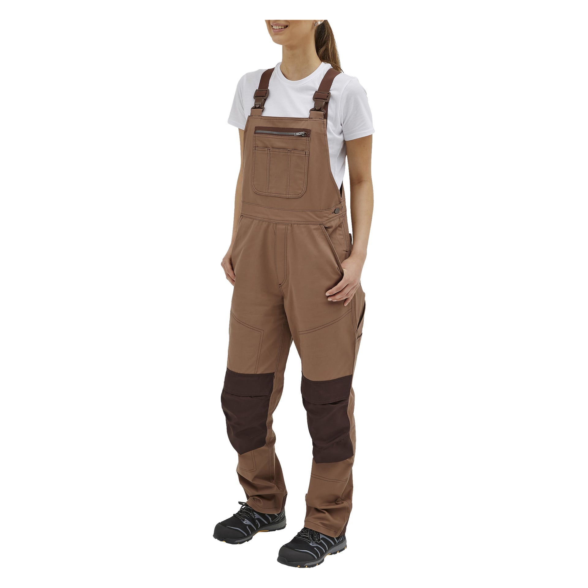En person står lätt vinklad, händerna i fickorna på bruna Jill Women's overaller från Bluewear med kontrasterande mörka fläckar på knäna, bära över en vit T-shirt och kompletteras av svarta arbetsskor.