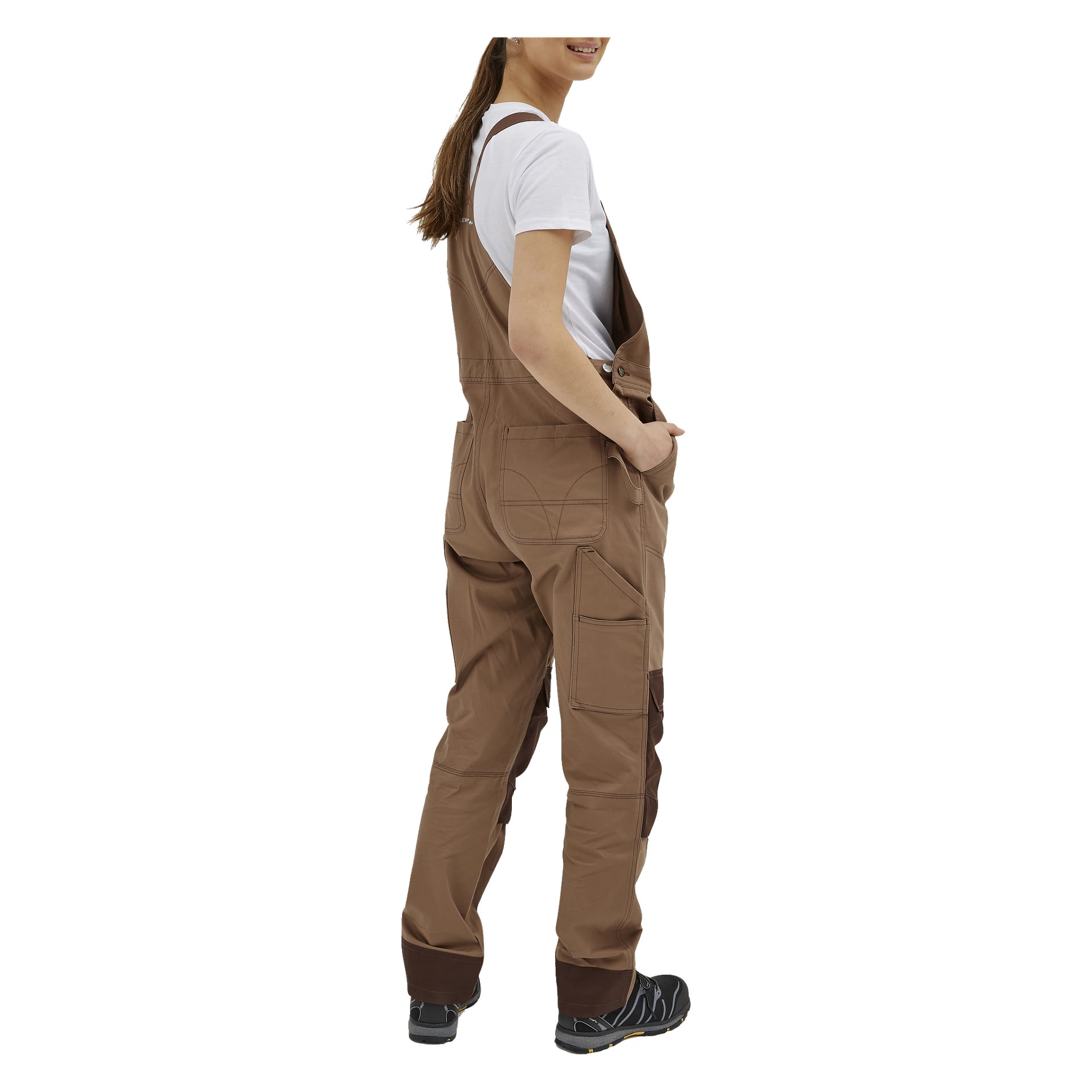 En person står litt vendt bort, kledd i Bluewear Women's Jill-overalls—brune kjeledresser med store lommer og mørke knelapper—sammen med en hvit t-skjorte og svarte sko. Håret deres er trukket tilbake i en hestehale.