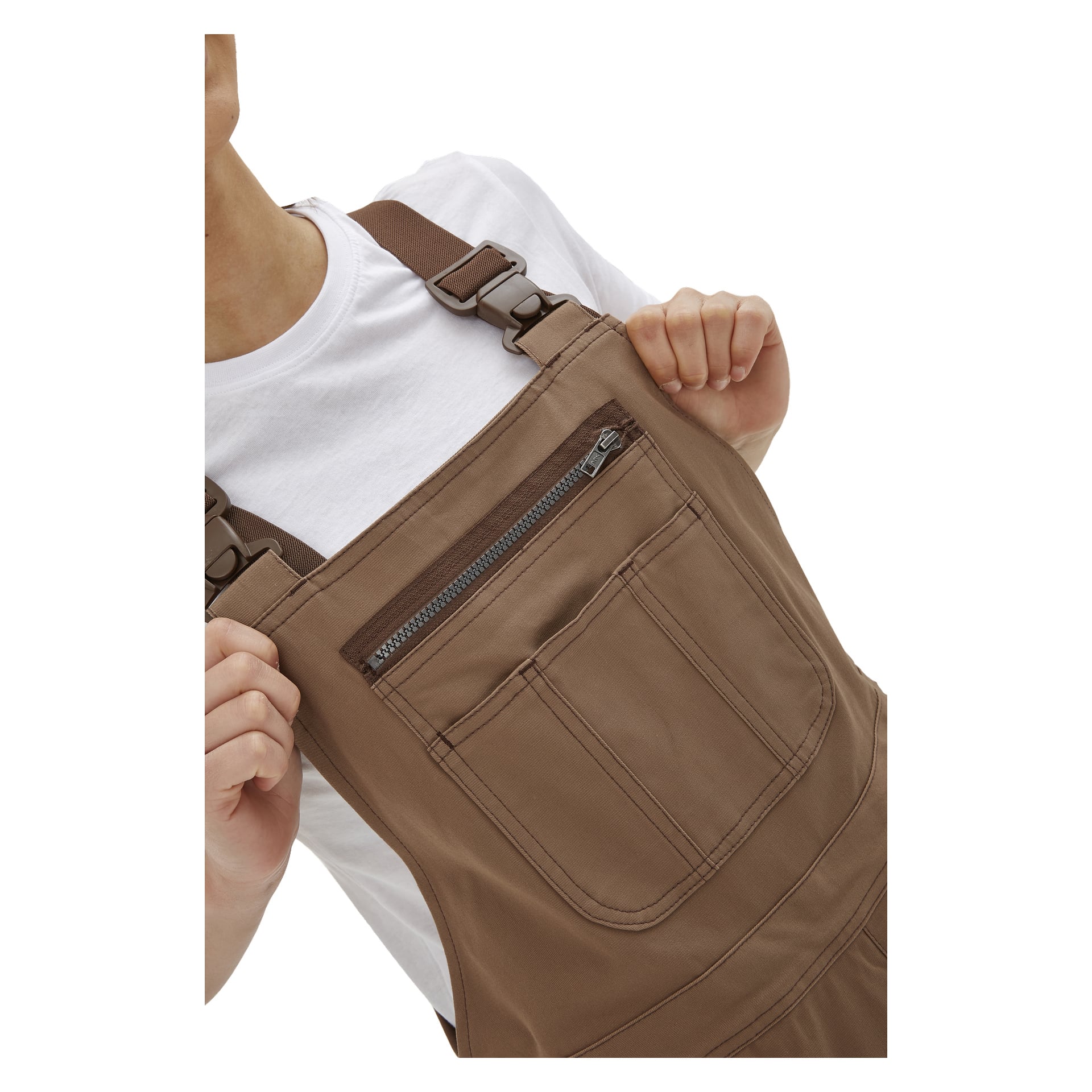 Utsikt fra skulder til midje av noen som har på seg brune Bluewear Women's Jill-kjeledresser med glidelåslomme foran, stylet over en hvit t-skjorte. Personen holder fast i begge stroppene, og ansiktet forblir utenfor bildet.