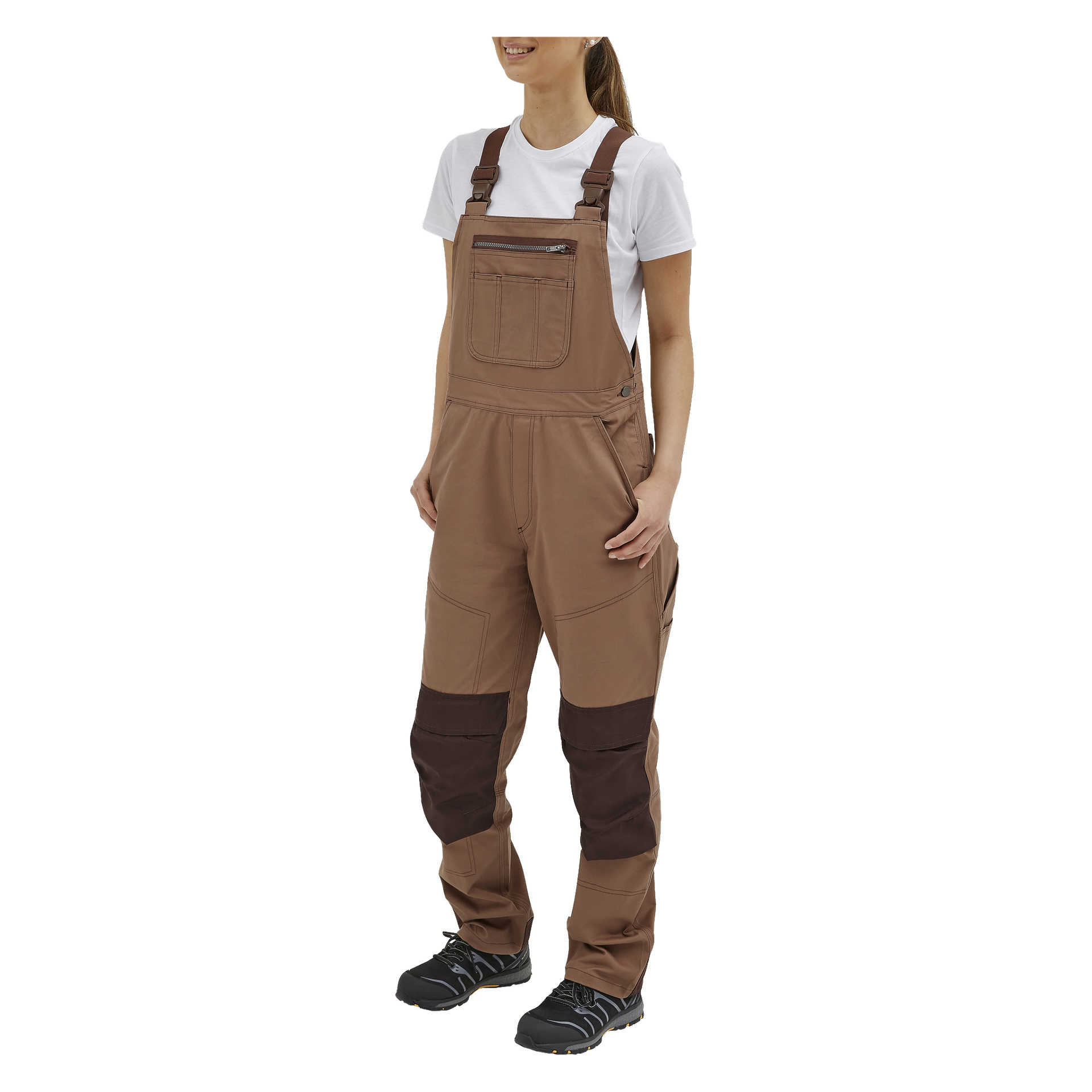Man trägt die Bluewear Women's Jill Overalls, darunter ein weißes T-Shirt und dunkelbraune Kniepatches. Sie stehen leicht zur Seite gedreht, beide Hände in den Taschen und tragen schwarze Arbeitsschuhe.