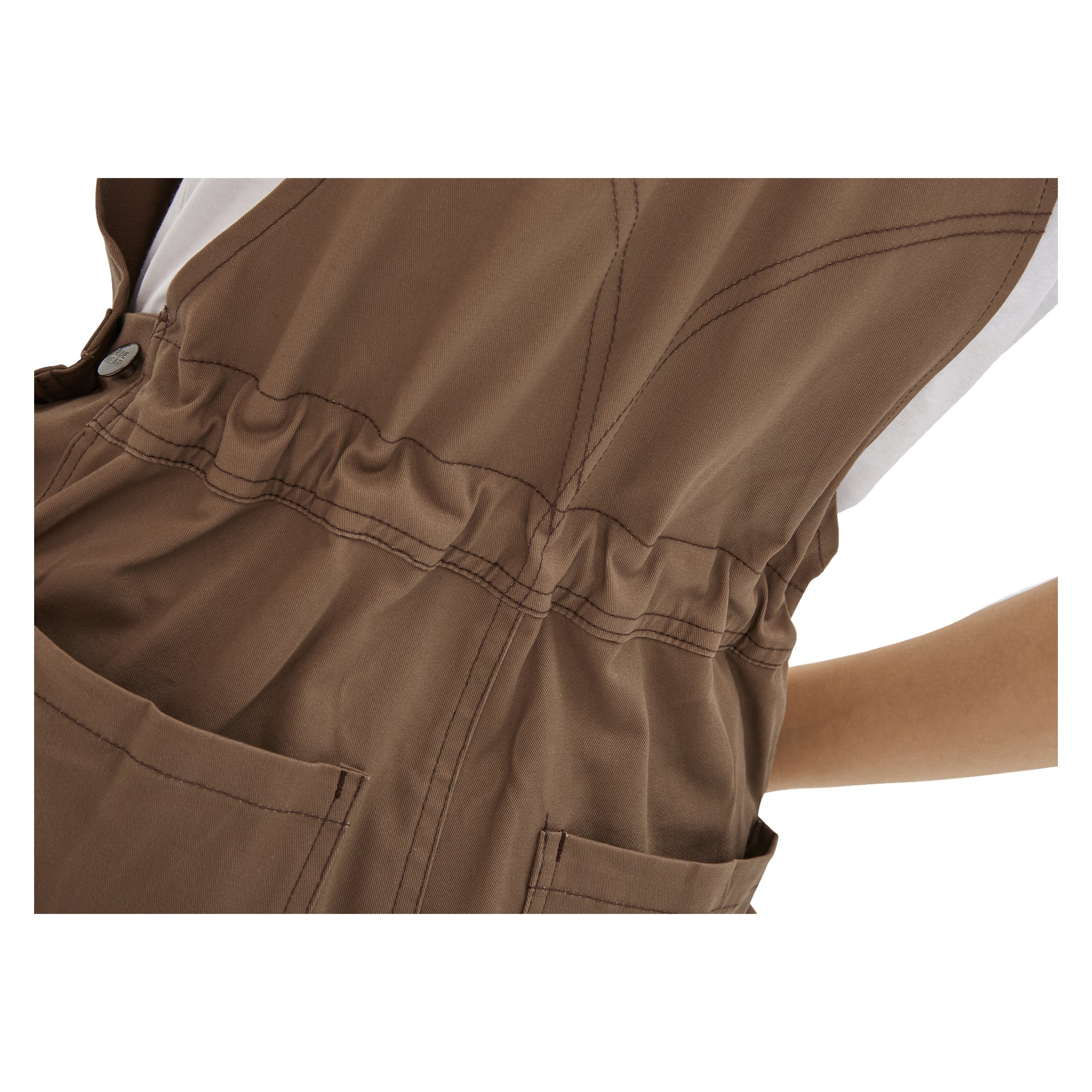 Eine Nahaufnahme zeigt jemanden, der die braunen Damenlatzhosen von Bluewear Jill trägt, mit zwei vorderen Taschen, auffälligen Nähten und einem elastischen Bund. Ein Teil eines weißen Hemdes und der Arm des Trägers sind ebenfalls zu sehen.