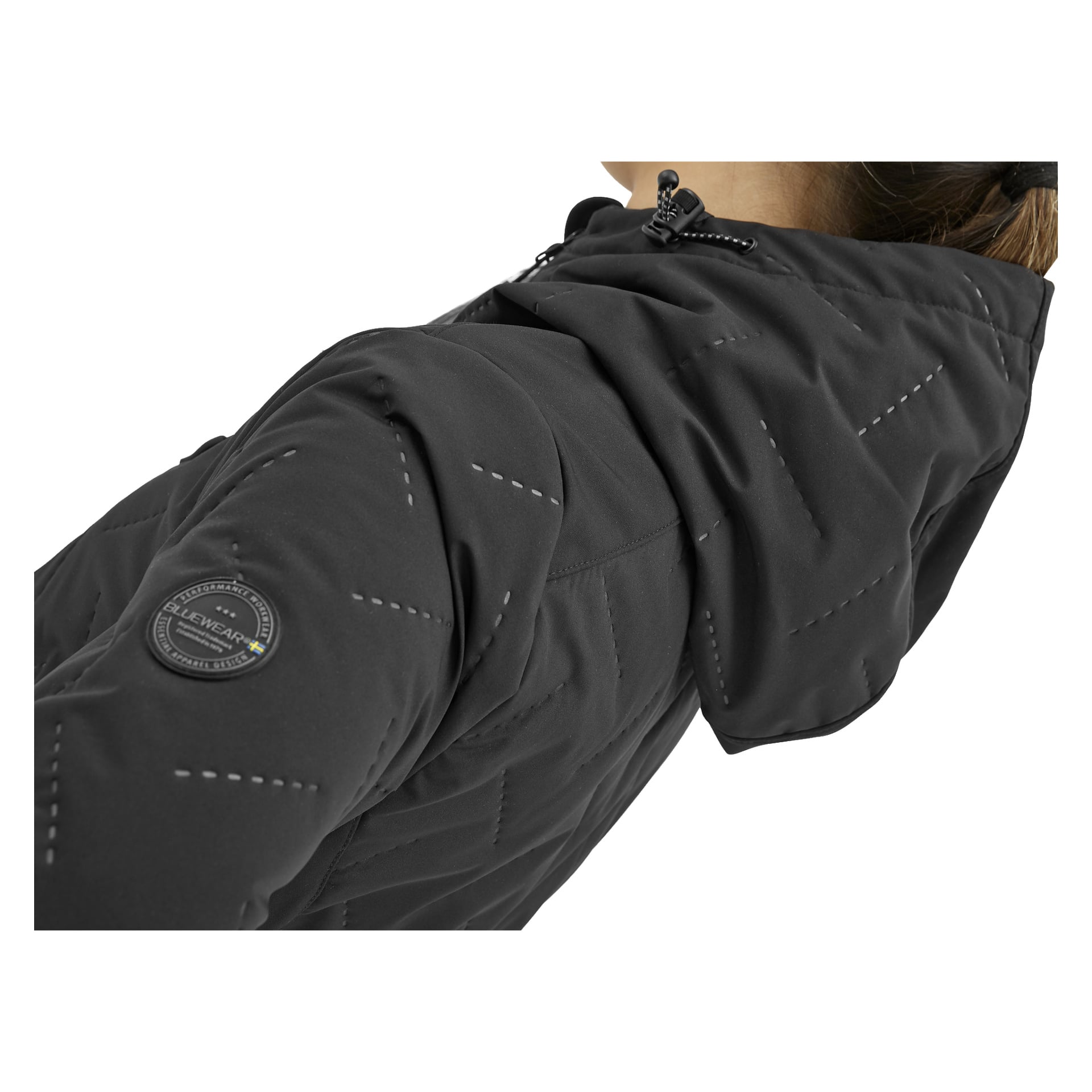 En närbild visar en person klädd i Bluewear Jenny softshelljacka för kvinnor, i svart. Jackan har diagonala quiltade mönster, en huva och en rund lapp på överarmen. Endast personens huvud och axlar är delvis synliga.