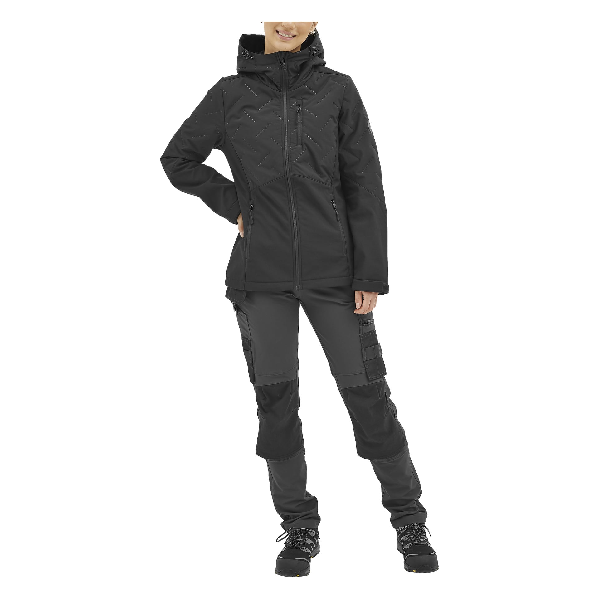 En person står framåt, klädd i Bluewear Jennys lättviktsvadderade softshell-jacka för kvinnor i svart, tillsammans med mörka cargobyxor med flera fickor och svarta skor. Deras vänstra hand är på höften och de ler svagt.