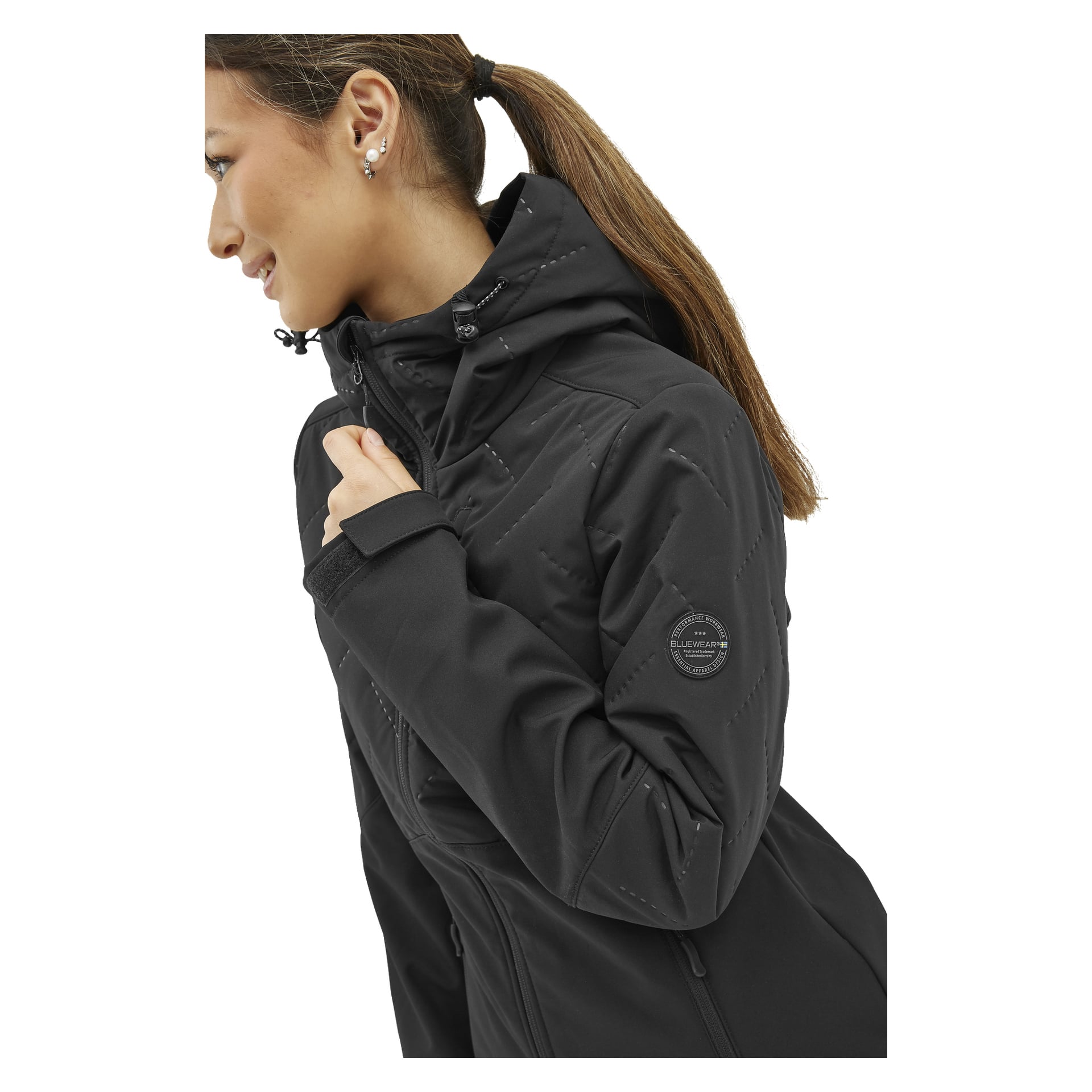 Sivulta esillä oleva nainen esitteli Bluewear Lightly -pehmeäkuorista naisten softshell-takkia Jennyä, jossa on musta vetoketjullinen kuvio ja huppuri. Hän tarttuu kaulukseen yhdellä kädellä, korostaen ylähihan pyöreää paikkaa ja erottuvia ompeleita. Hänen hiuksensa on sidottu poninhännälle ja hänellä on korvakorut.