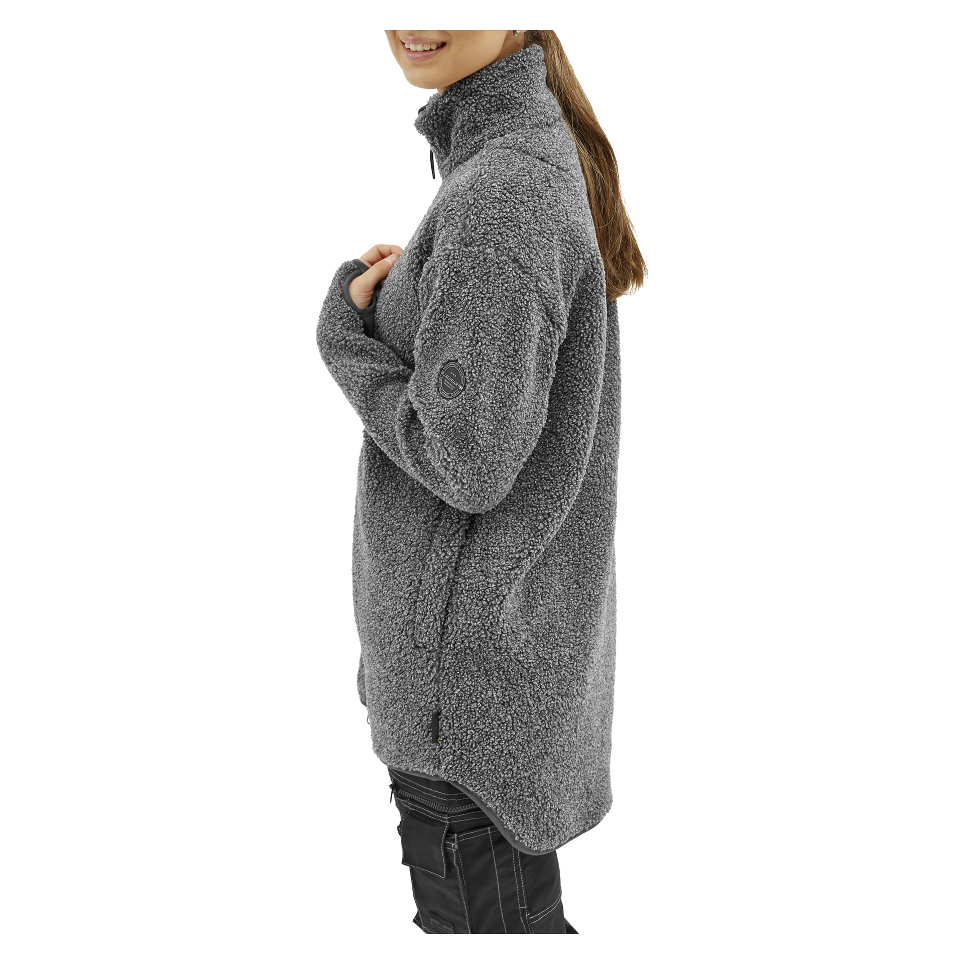 Eine lächelnde Frau mit langen, nach hinten gebundenen braunen Haaren steht im Profil, trägt eine graue Bluewear Vivienne Fleecejacke mit strukturiertem Stoff, Reißverschluss vorne, hohem Kragen und einem runden Patch am oberen Ärmel.
