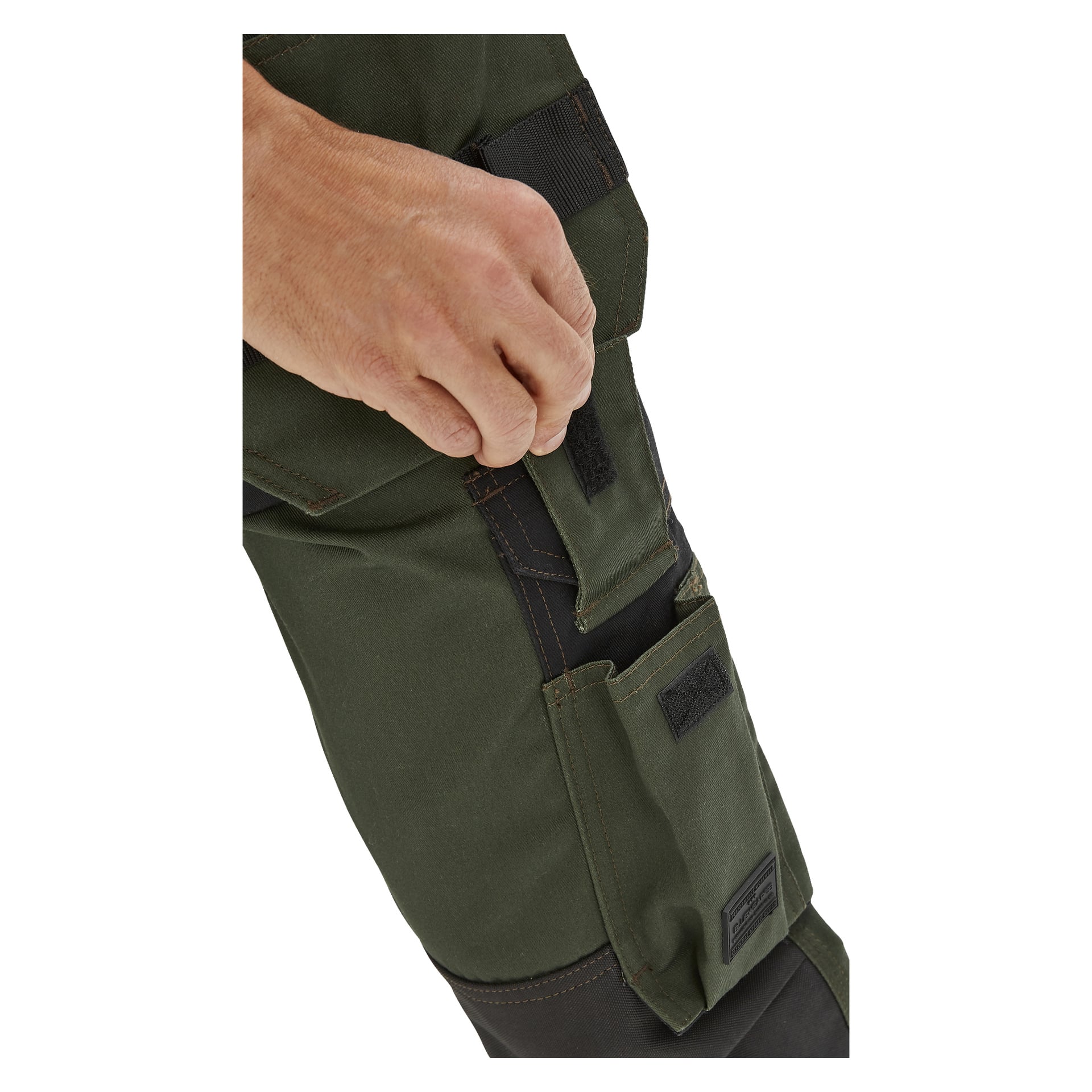 En hand öppnar en kardborreficka på Bluewear herrbyxor Mr Green. Byxorna är mörkgröna och svarta med flera fickor och förstärkta sömmar.