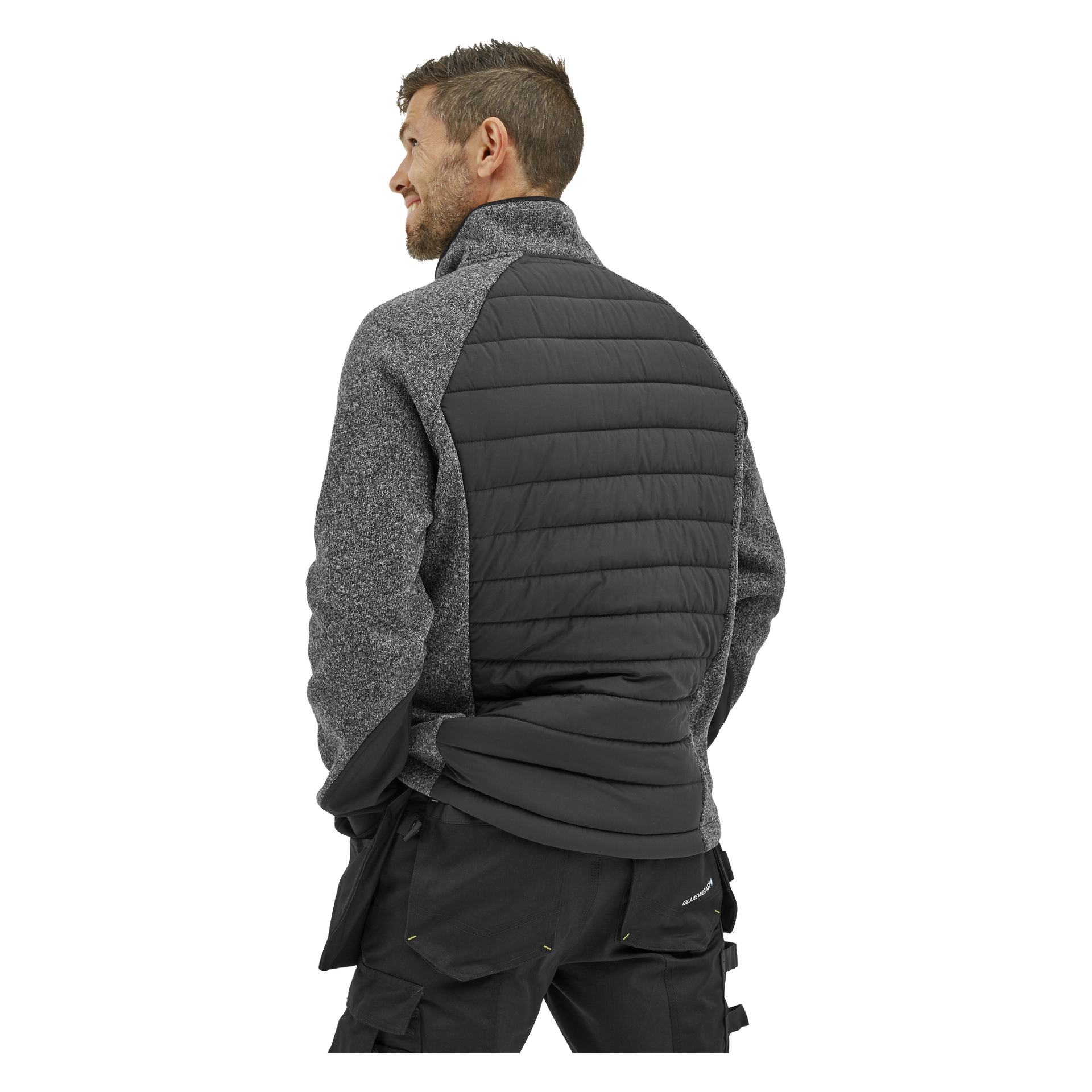 Mężczyzna z krótkimi brązowymi włosami stoi tyłem do kamery, ubrany w "Bluewear Men's hybrid jacket Hilmer" – szaro-czarną kurtkę z pikowanym panelem na plecach oraz czarne spodnie.