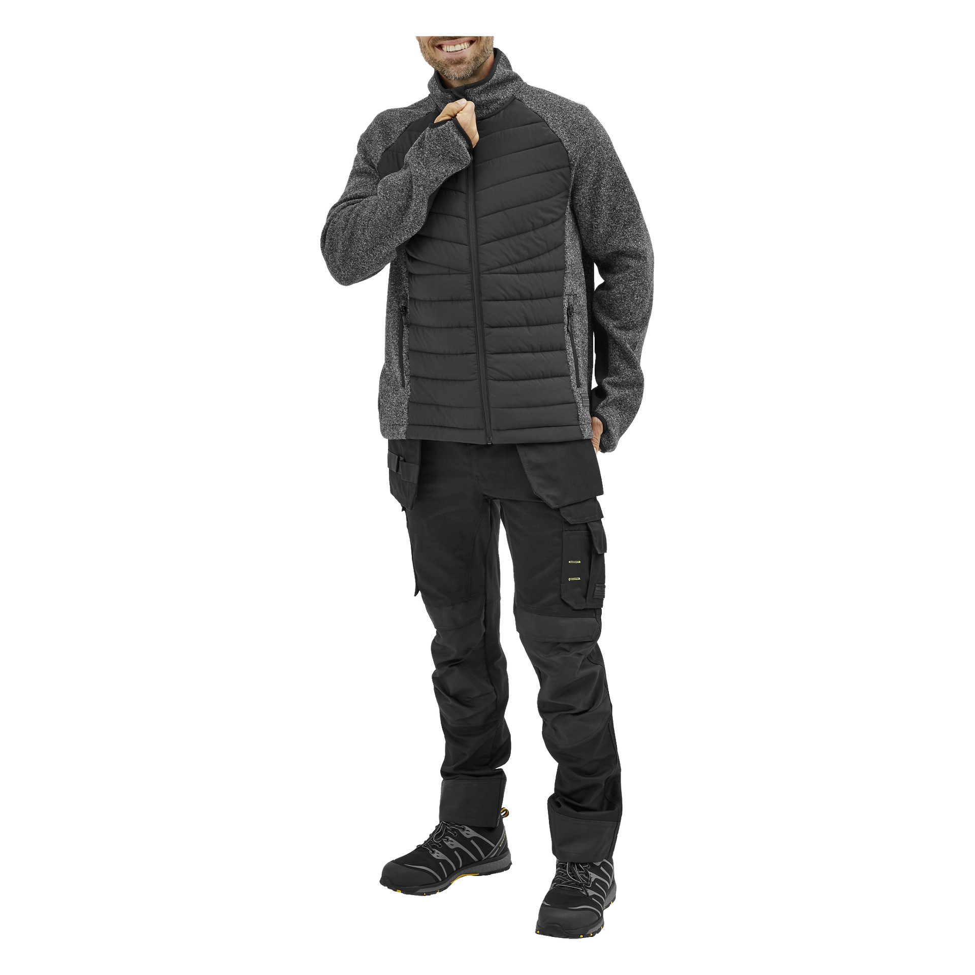 Mężczyzna się uśmiecha, ubrany w "Bluewear Men's hybrid jacket Hilmer" w czerni i szarości, w czarne spodnie cargo i buty trekkingowe. Prawa ręka spoczywa na kołnierzu kurtki; widać tylko część twarzy.