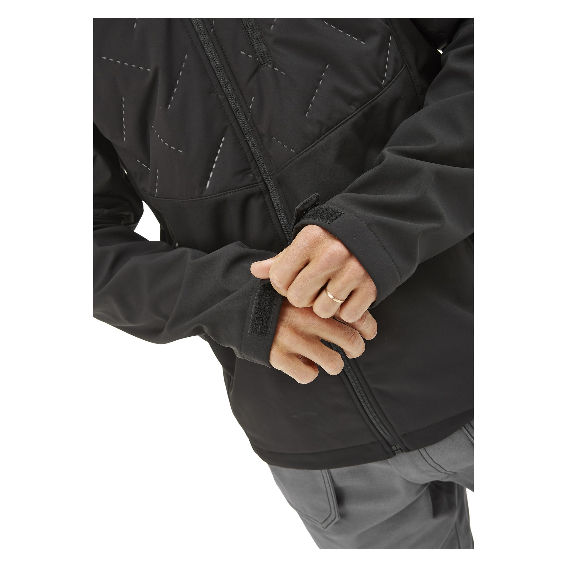 En person klädd i Bluewear Lätt vadderad herrsoftshelljacka Jerry och grå byxor justerar handledsremmen på ärmen. Endast deras överkropp och händer kan ses, och en guldring syns på deras vänstra hand.