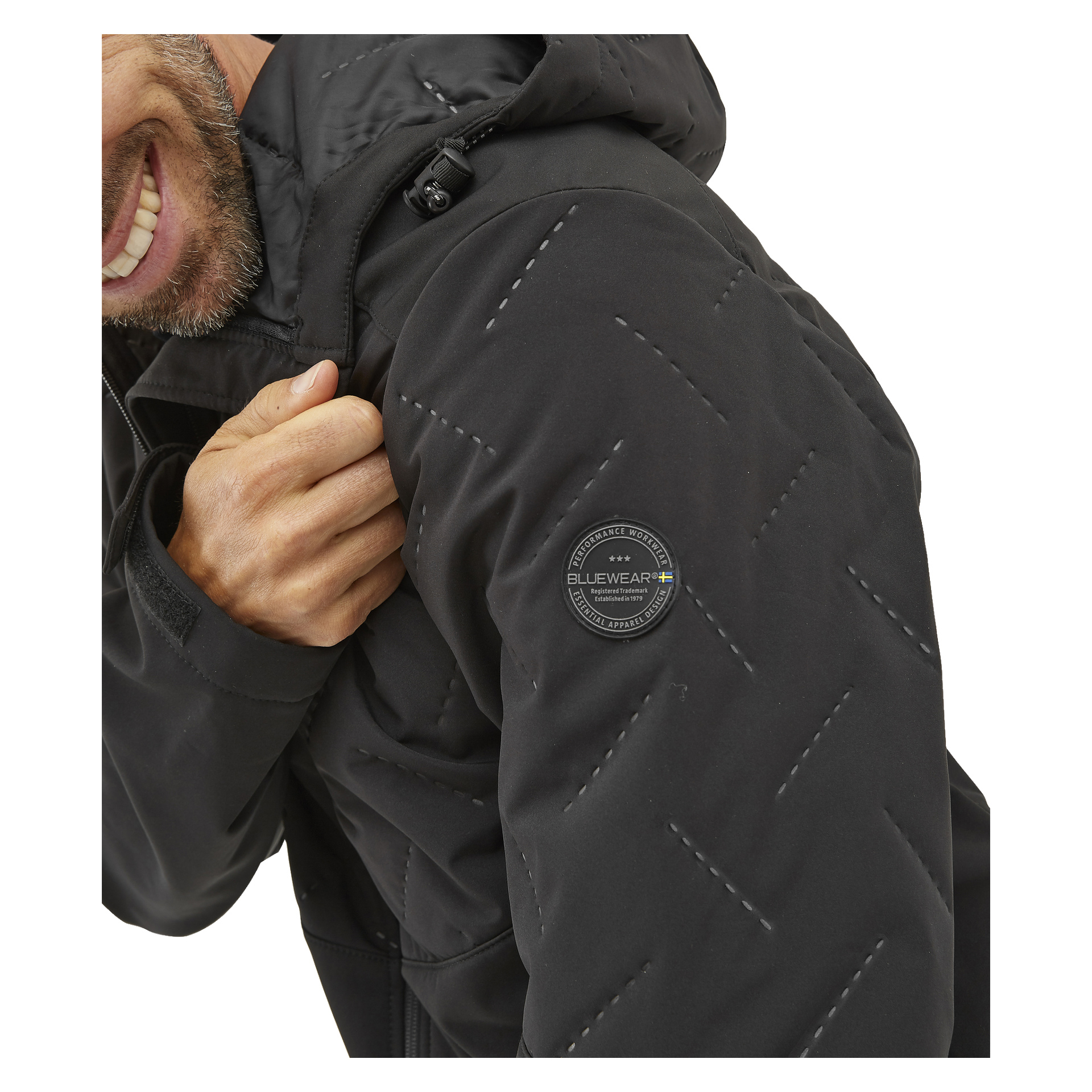 Någon i en Bluewear lätt vadderad herrsoftshell-jacka Jerry, med den karakteristiska runda Bluewear-patchen på överärmen, visas leende. Endast den nedre halvan av ansiktet och en del av överkroppen kan ses.