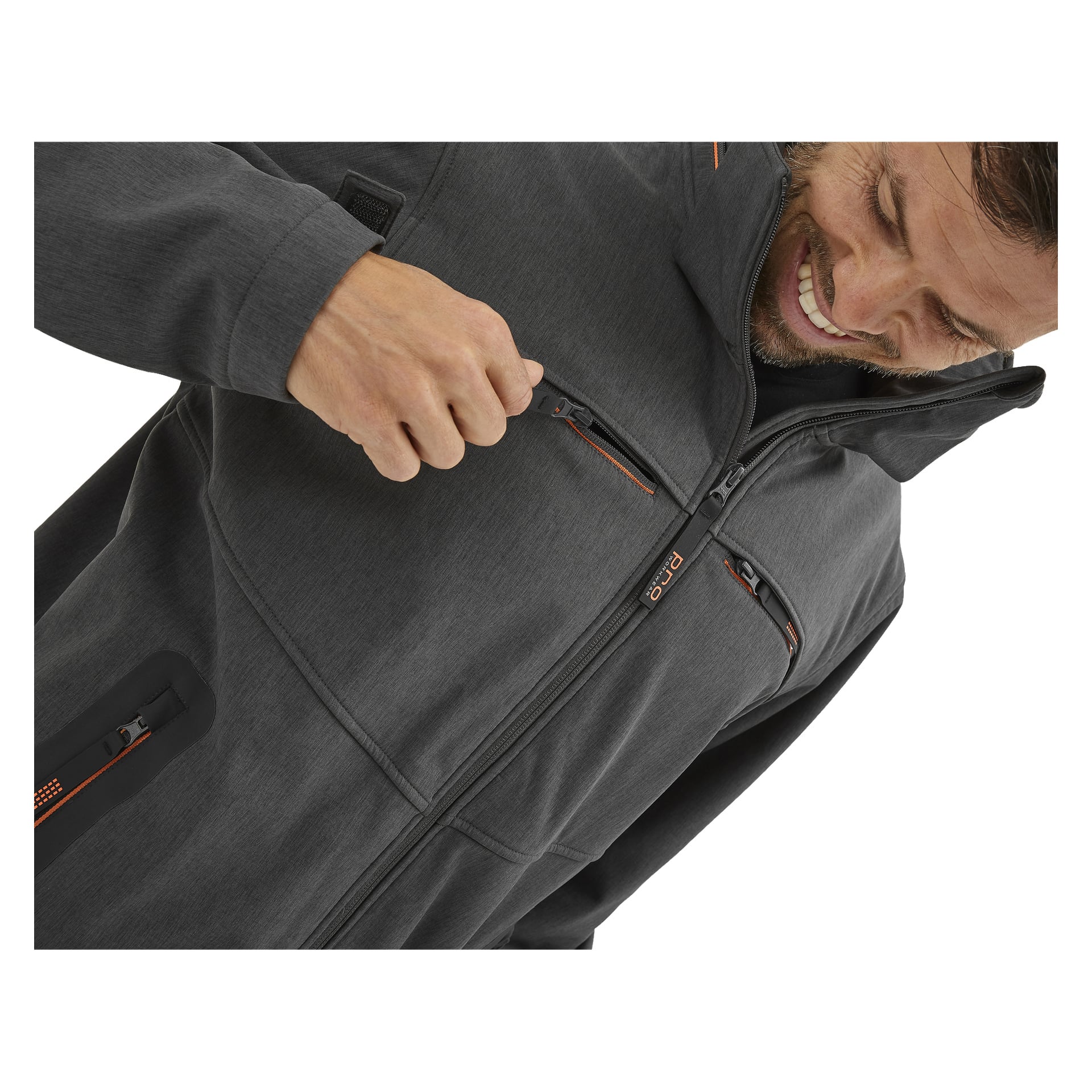En person ler när de öppnar bröstfickan på Bluewear Pro Workwear Pontus softshell-jacka för herrar, som är mörkgrå med orange dragkedjedetaljer. Bilden visar bara deras överkropp.