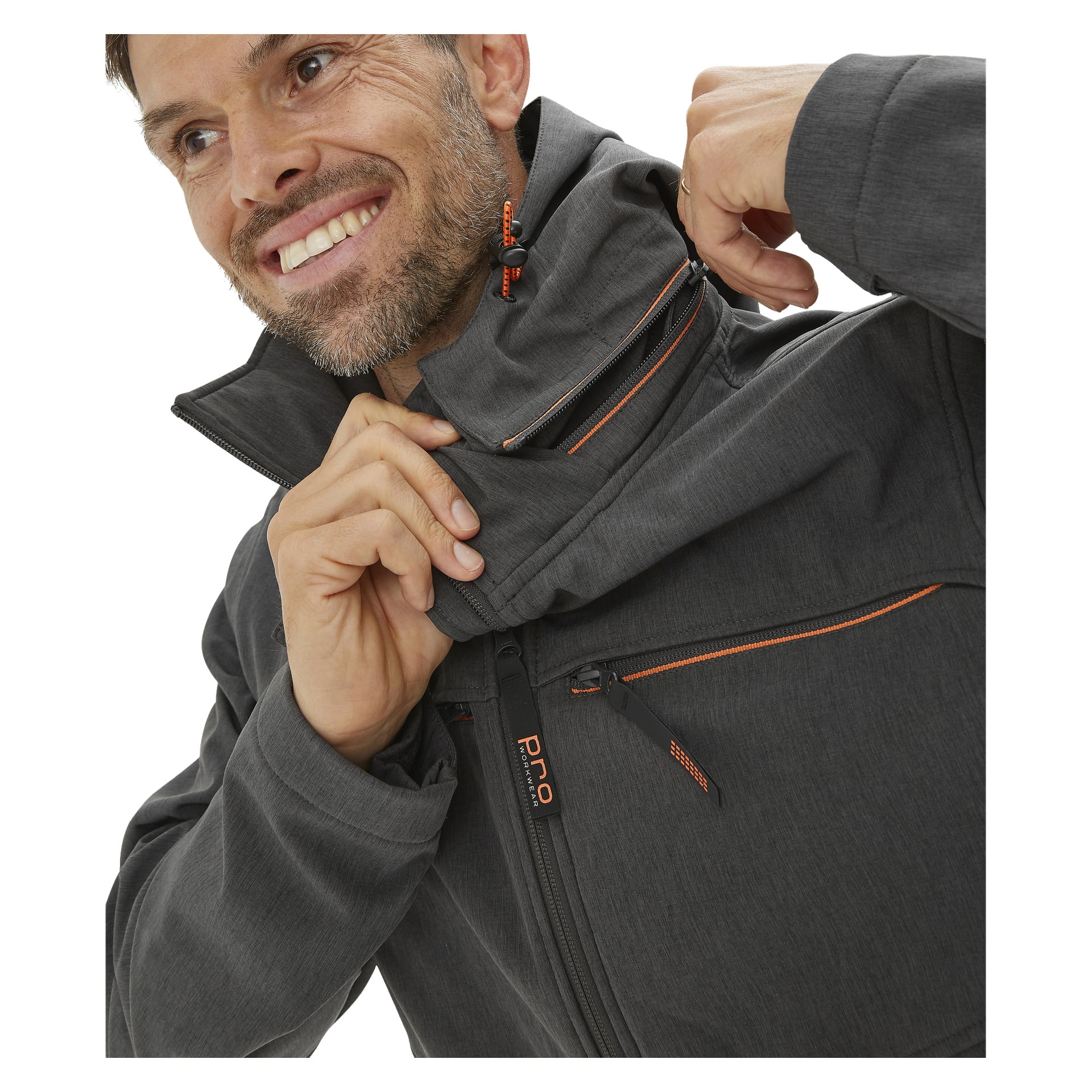 En man bär Bluewear Pro Workwear Men's Softshell Jacket Pontus i mörkgrått med orange dragkedjedetaljer. Han ler, lyfter kragen med ena handen och håller en dragkedja nära halsen med den andra.
