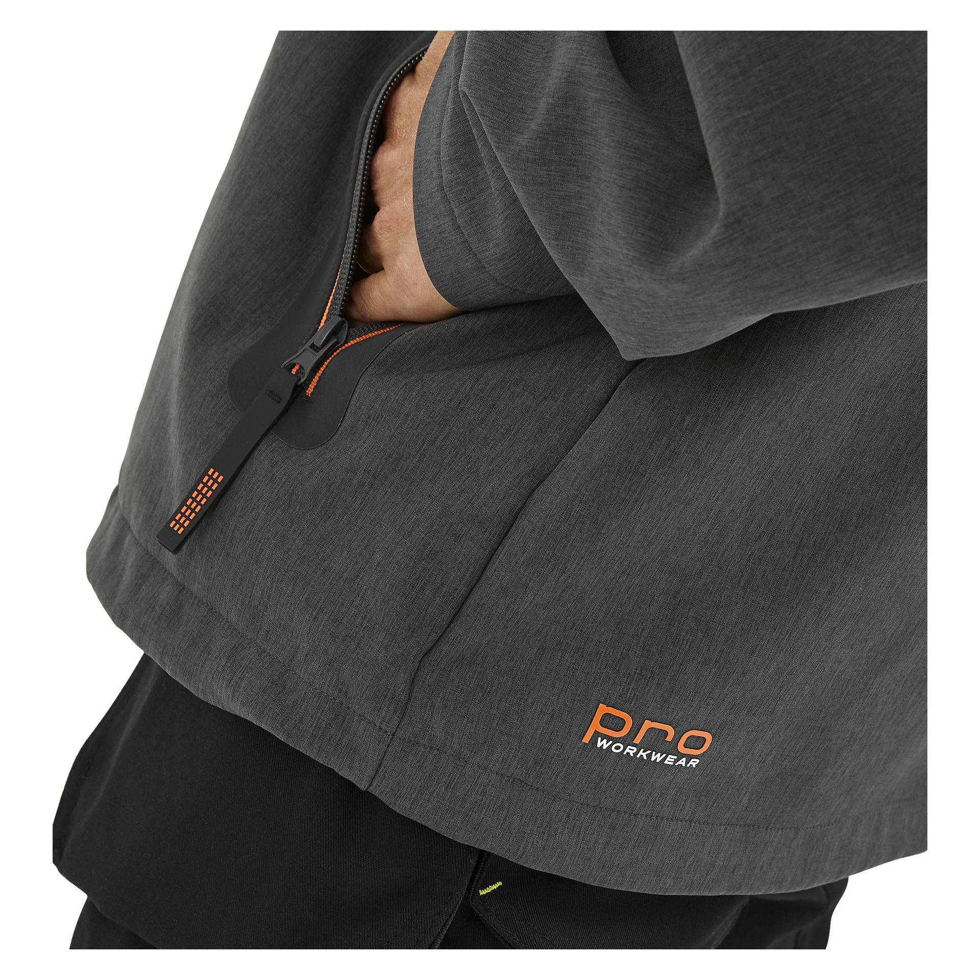En person visas från midjan och nedåt bär Pontus herrsoftshelljacka från Bluewear Pro Workwear, som är mörkgrå med orange logga och dragkedjedetaljer. Deras hand vilar i fickan, och endast nedre delen av överkroppen och en del av en arm syns.