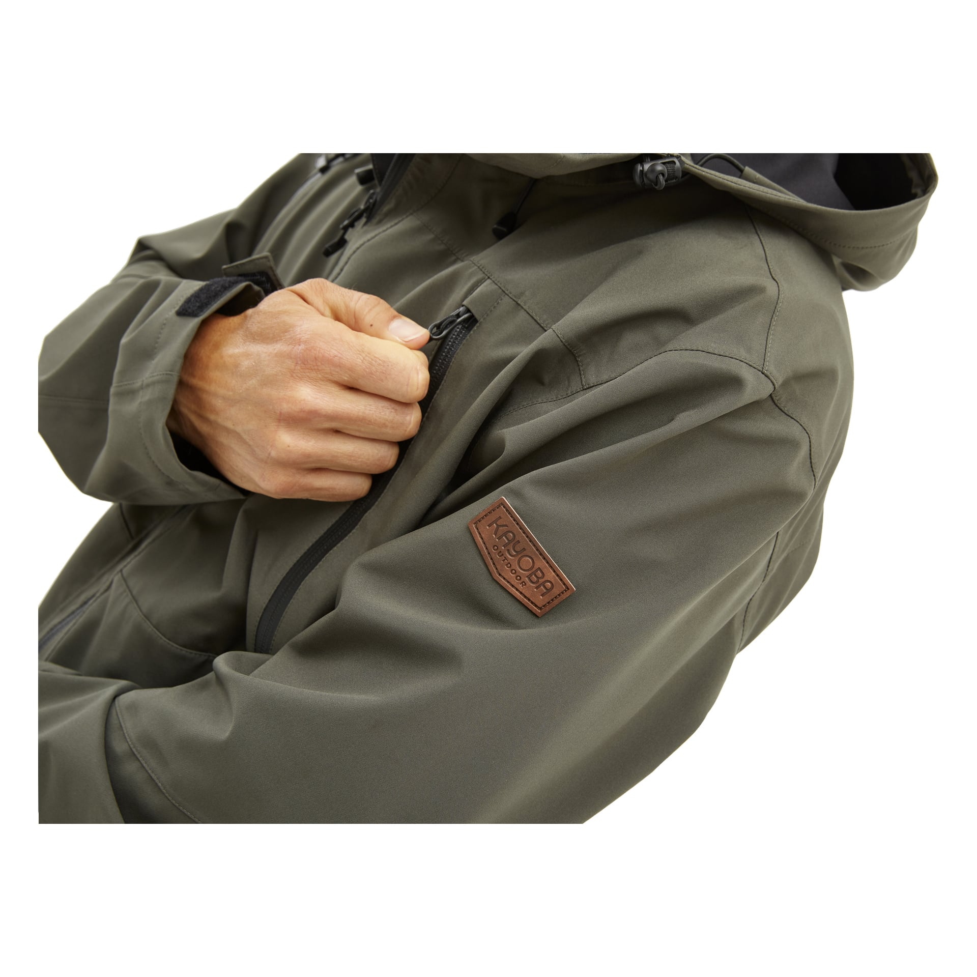 En person klädd i den olivgröna Kayoba Outdoor Men's Helag-jackan, som har ett Kryptek-märke på sig, drar ner dragkedjan på bröstfickan. Bilden visar bara deras överkropp och en hand.