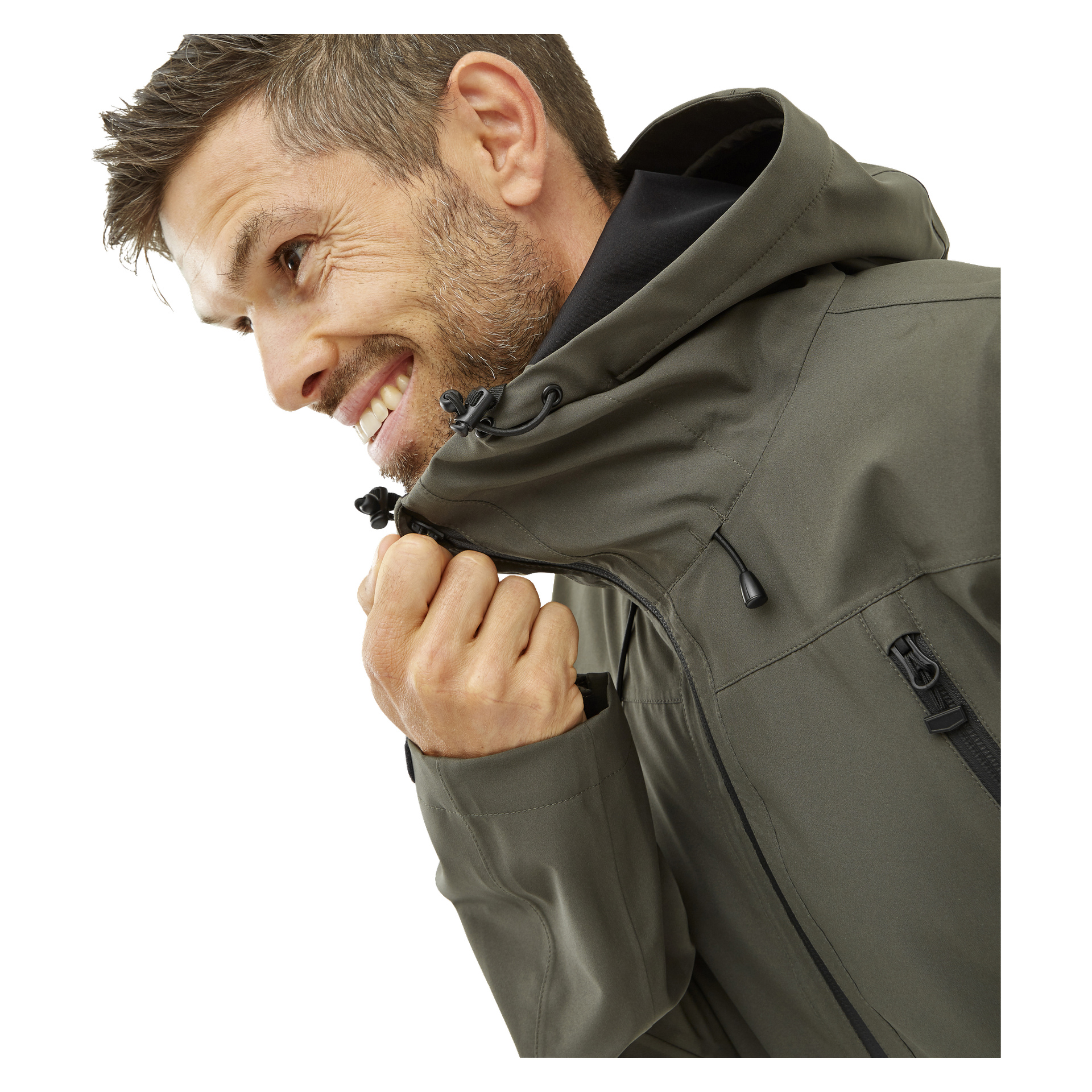 En man med kort brunt hår och ansiktshår ler när han drar igen en olivgrön Kayoba Outdoor Men's Shell Jacket Helag, som har svarta dragkedjor och snören.