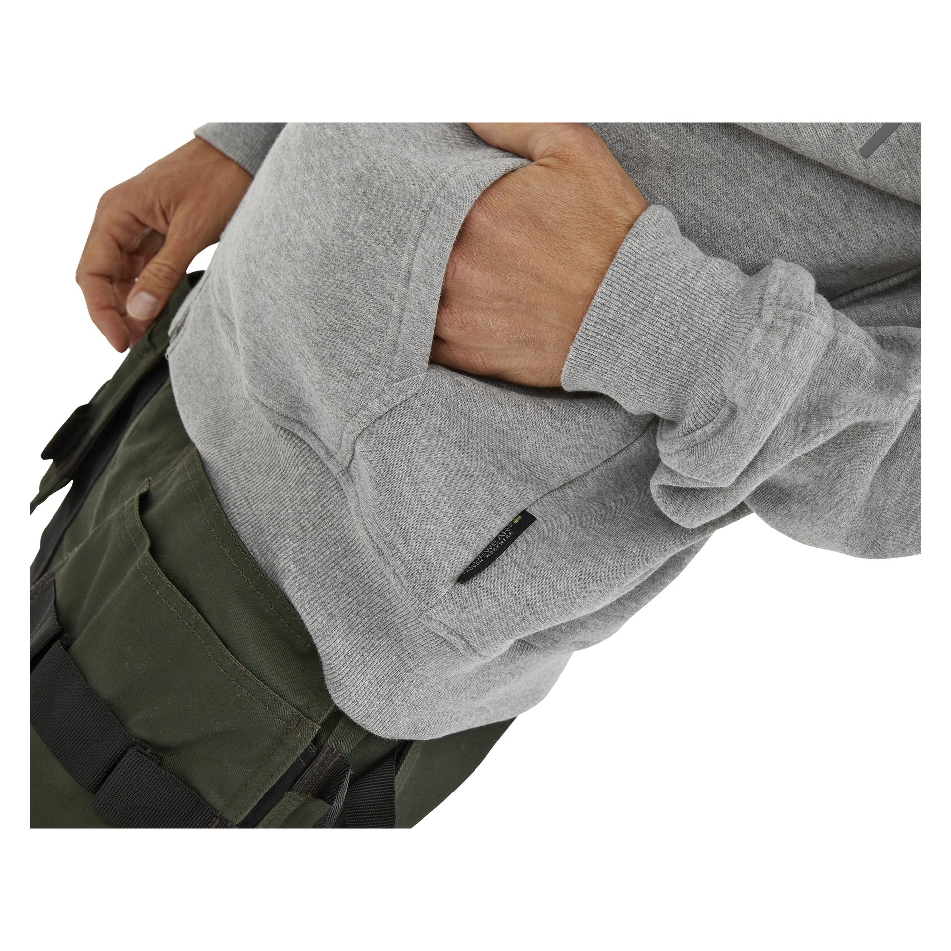 Personen i olivgröna byxor och en ljusgrå sweatshirt står med ena handen i sweatshirtens främre ficka, vilket visar överkropp och lår.