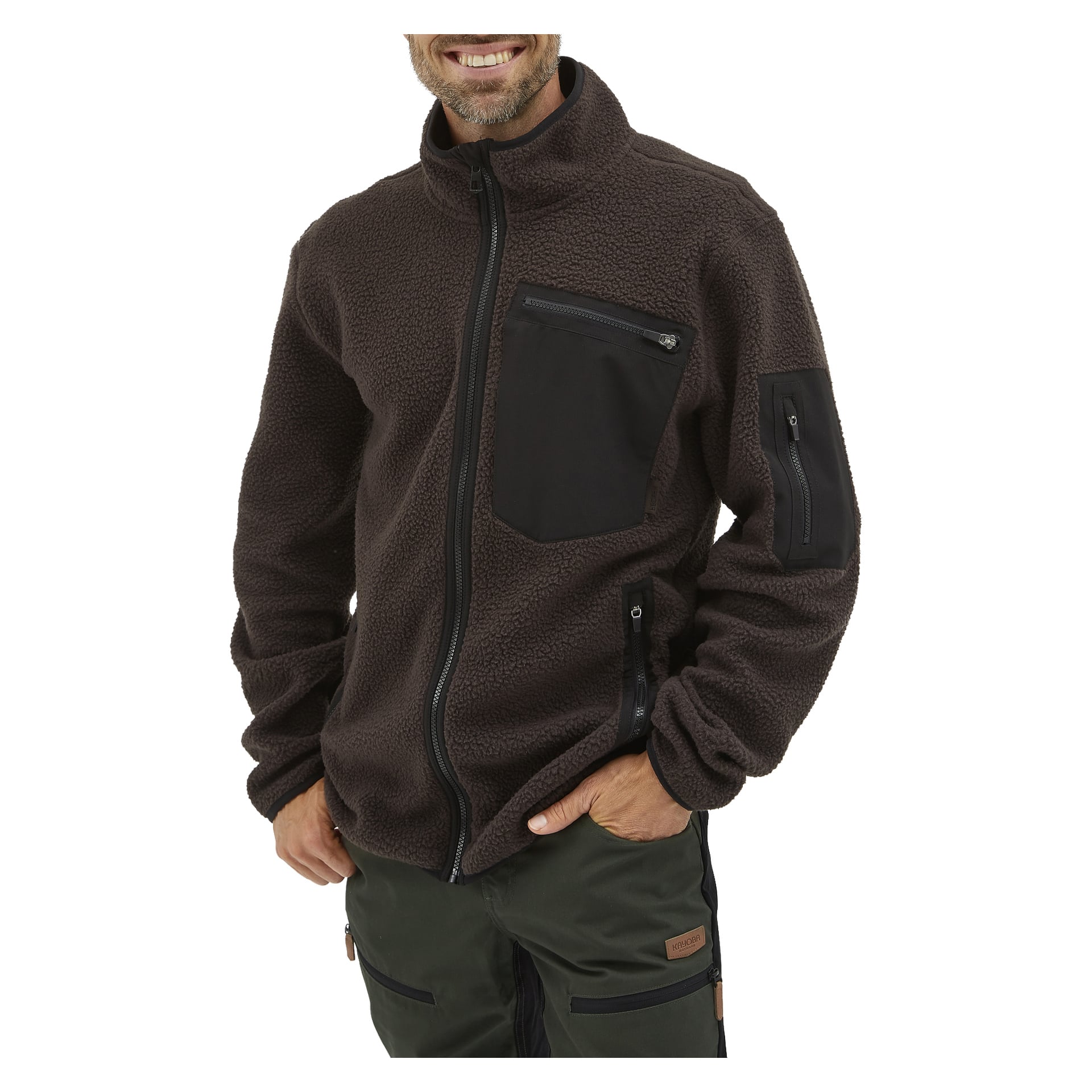 En person klädd i Kayoba Outdoor Men's pile jacket Ljusdal, en mörkbrun fleece-zip-up med svarta fickor med dragkedja på bröstet, vänster ärm och båda sidorna, tillsammans med mörkgröna byxor. Personen ler och har ena handen i fickan.
