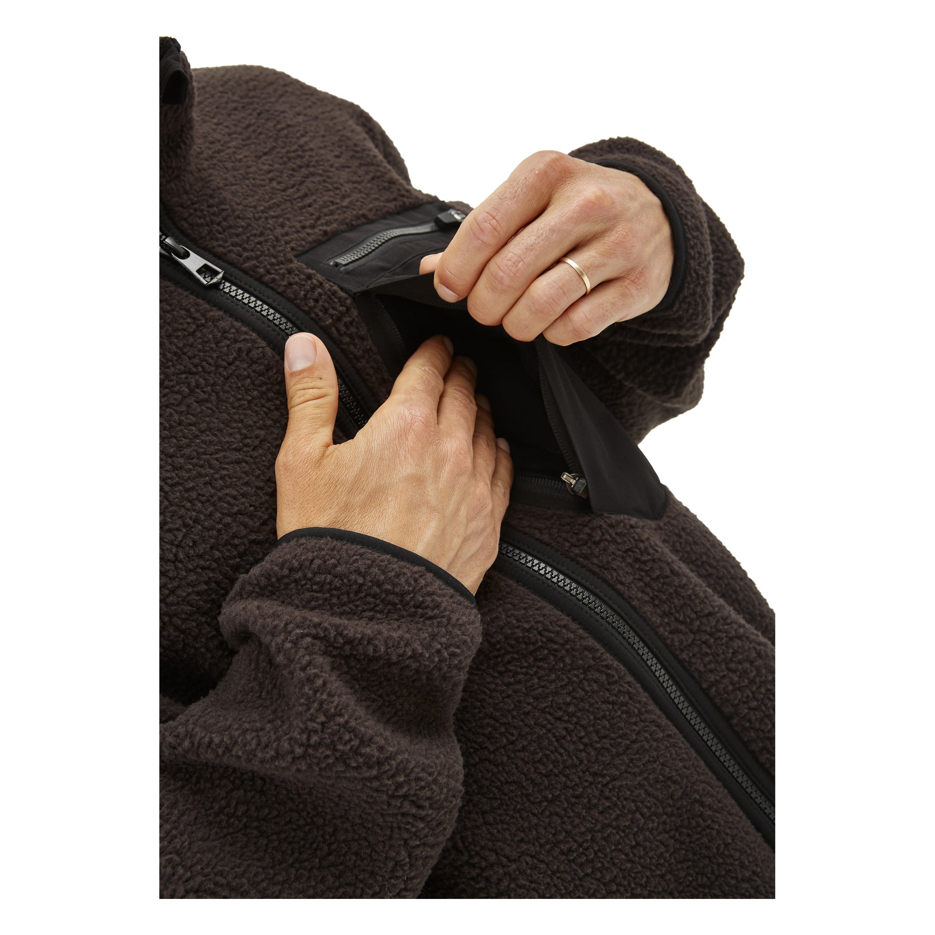 Någon klädd i en mörkbrun Kayoba Outdoor Men's pile-jacka Ljusdal använder ena handen för att dra ner dragkedjan och öppna den svarta bröstfickan, medan den andra handen sträcker sig in. En guldring kan ses på ett av deras fingrar.