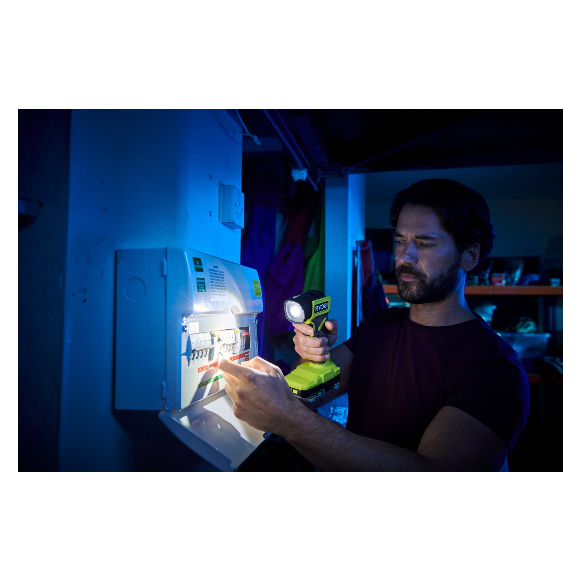En mann lyser med en grønn Ryobi One+ Torch RLF18-0 lommelykt inn i et elektrisk panel mens han peker på bryterne, og jobber i et svakt opplyst miljø med blåaktige toner som indikerer svakt lys eller strømbrudd.