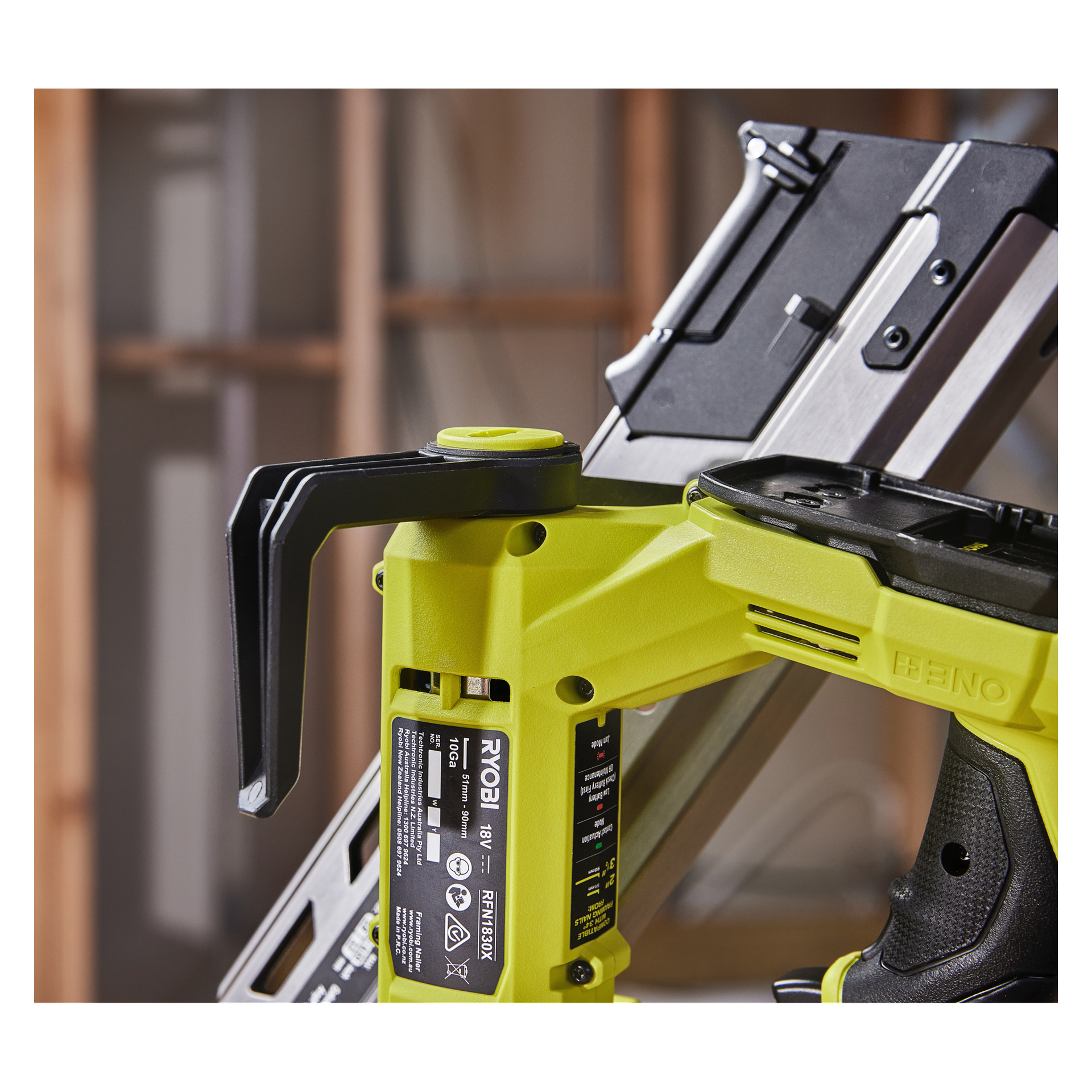 Lähikuva keltaisesta Ryobi One+ HP -akkukäyttöisestä naulapistoolista RFN1834X-0, jossa on musta kahva ja näkyvät etiketit, sumeaa taustaa vasten, jossa näkyy puupalkkeja ja metalliesine.