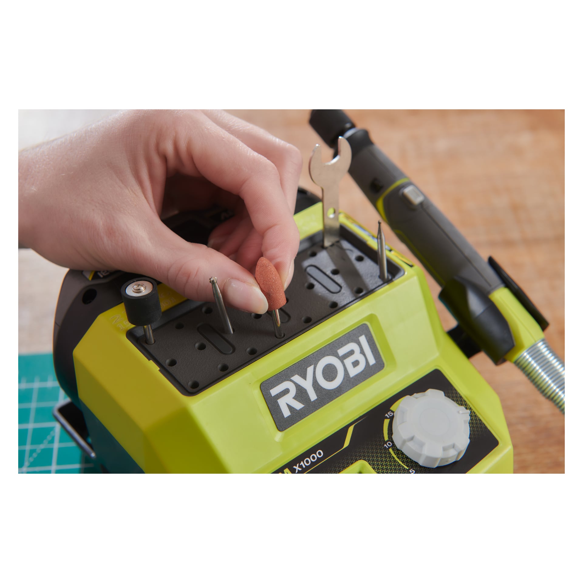 En hand för in en orange slipbit i en spår på en grön Ryobi One+ Multitool RRTS18-0A35-organisatör. Ytterligare bitar, en skiftnyckel och en del av verktyget vilar på en träyta i närheten.