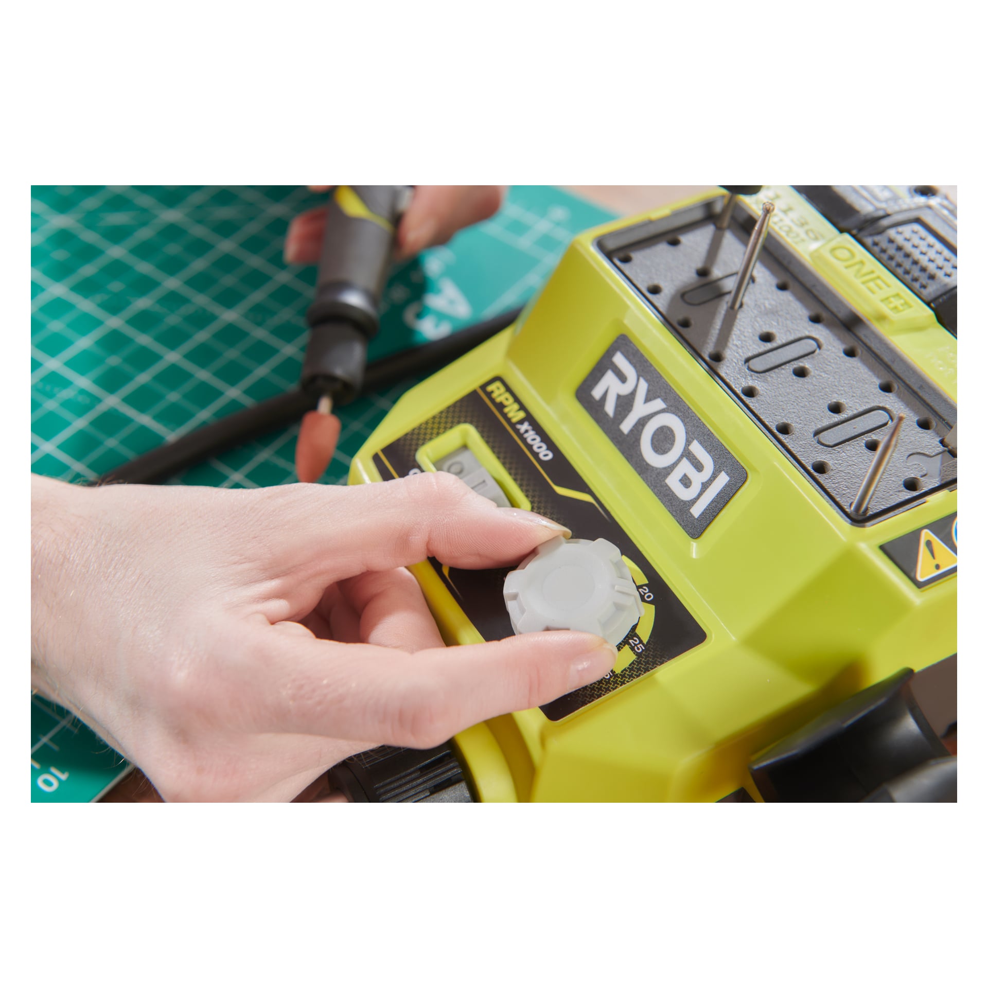 En hand vrider snabbratten på en gul Ryobi One+ Multitool RRTS18-0A35, medan en annan hand stabiliserar ett verktyg med en snurrande borr, allt placerat på en grön skärmatta.