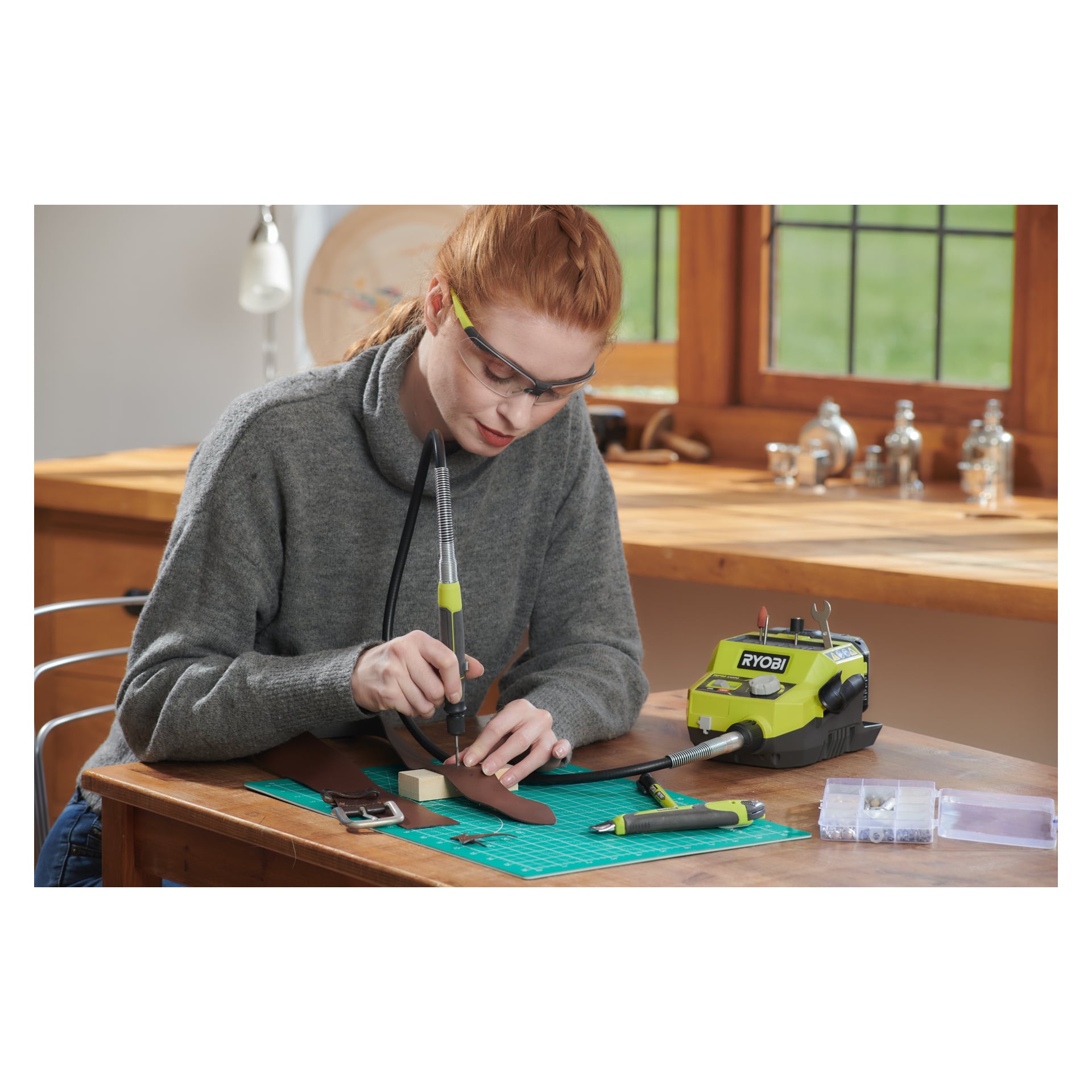 En person med skyddsglasögon använder en Ryobi One+ Multi-tool RRTS18-0A35 för att arbeta på en läderrem vid ett träbord. Skrivbordet innehåller ytterligare verktyg, inklusive ett annat grönt Ryobi One+-verktyg och en skärmatta, med solljus som strömmar in genom fönstren bakom dem.