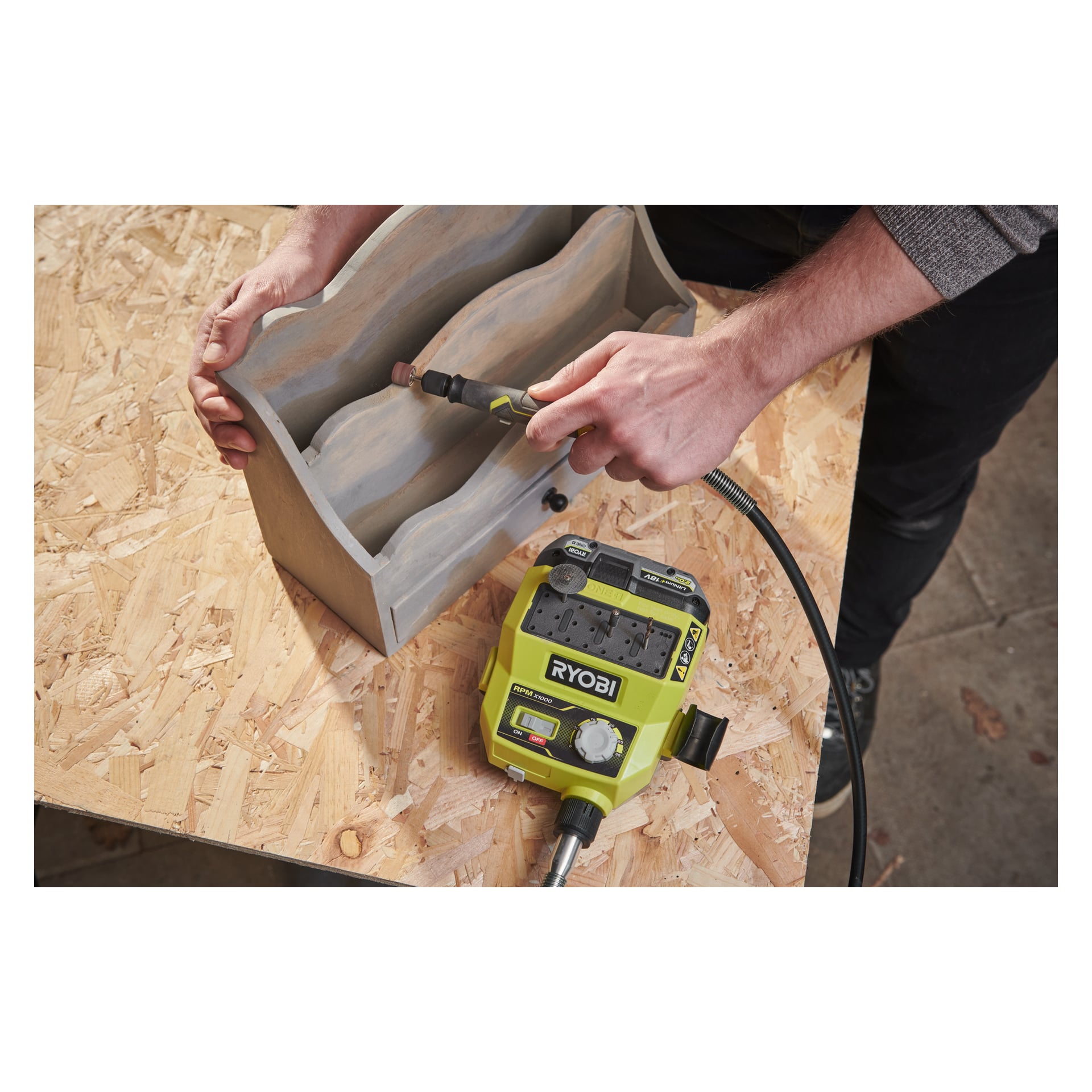Någon slipar insidan av en liten trähylla placerad på en arbetsbänk i plywood med hjälp av en Ryobi One+ Multitool RRTS18-0A35, som är kopplad till sin strömenhet via en flexibel sladd.