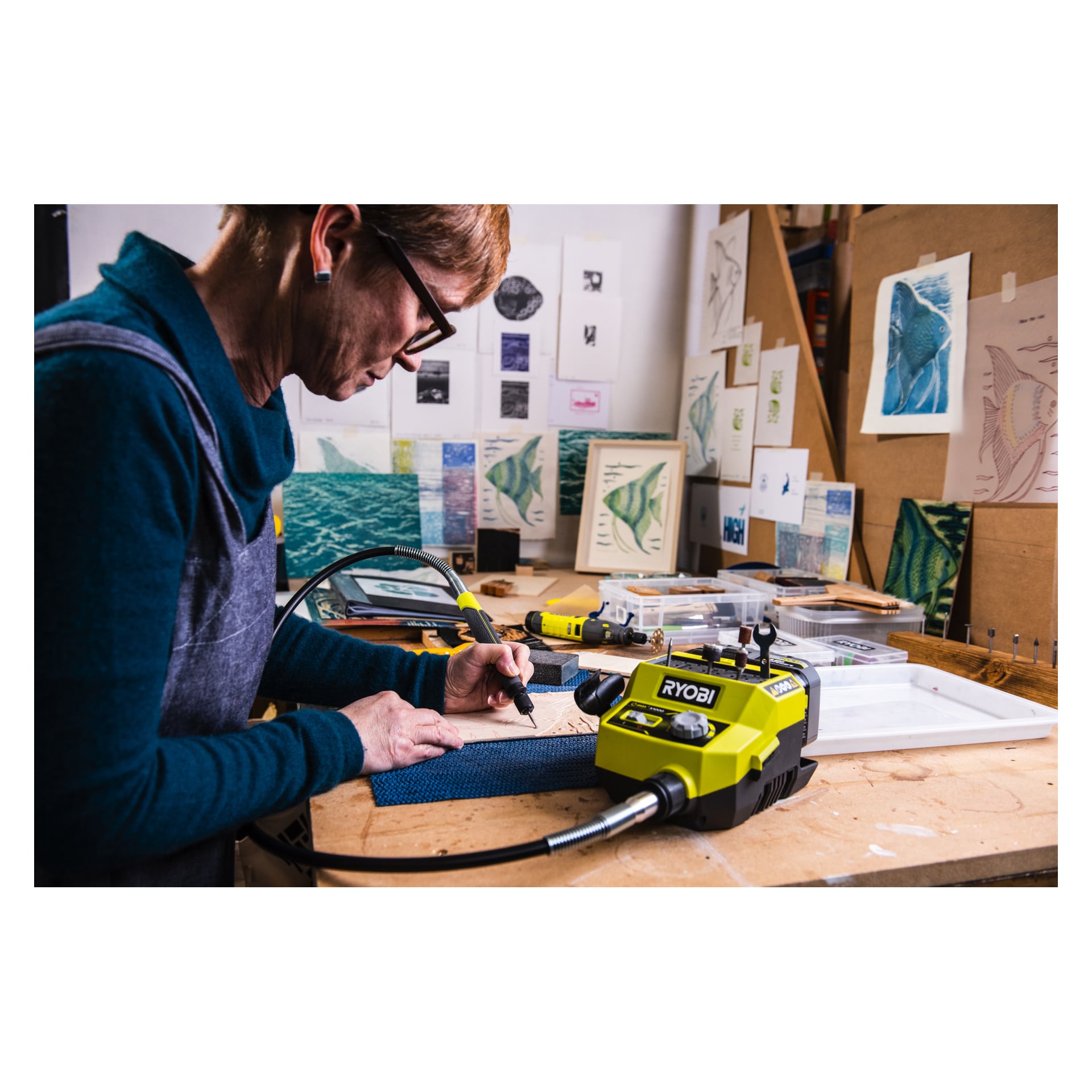 Någon arbetar vid ett trångt skrivbord och använder en Ryobi One+ Multitool RRTS18-0A35 på en liten materialbit. Arbetsplatsen har olika verktyg och fiskinspirerad konst, med tryck och skisser utställda på skrivbordet och väggen.