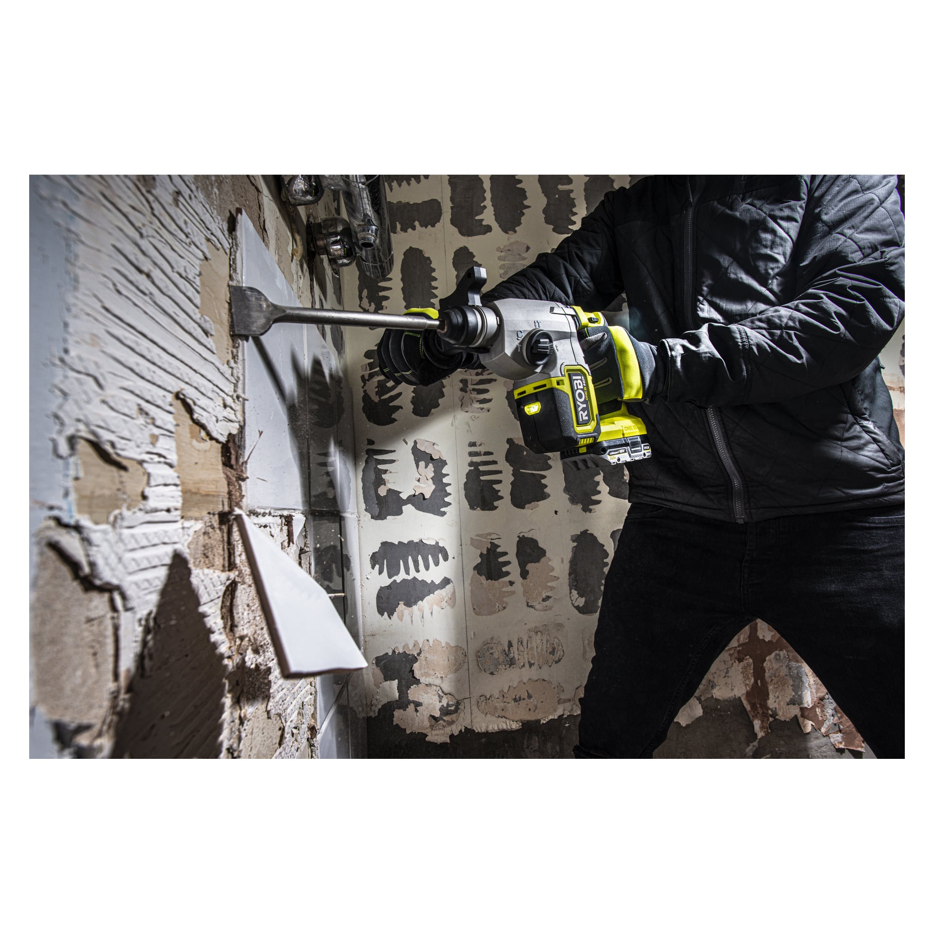En person i svart jacka använder en Ryobi One+ HP batteridriven hammarborr RSDS18X-0 med en mejselanordning för att flisa bort vita kakel från en vägg under rumsrenovering.