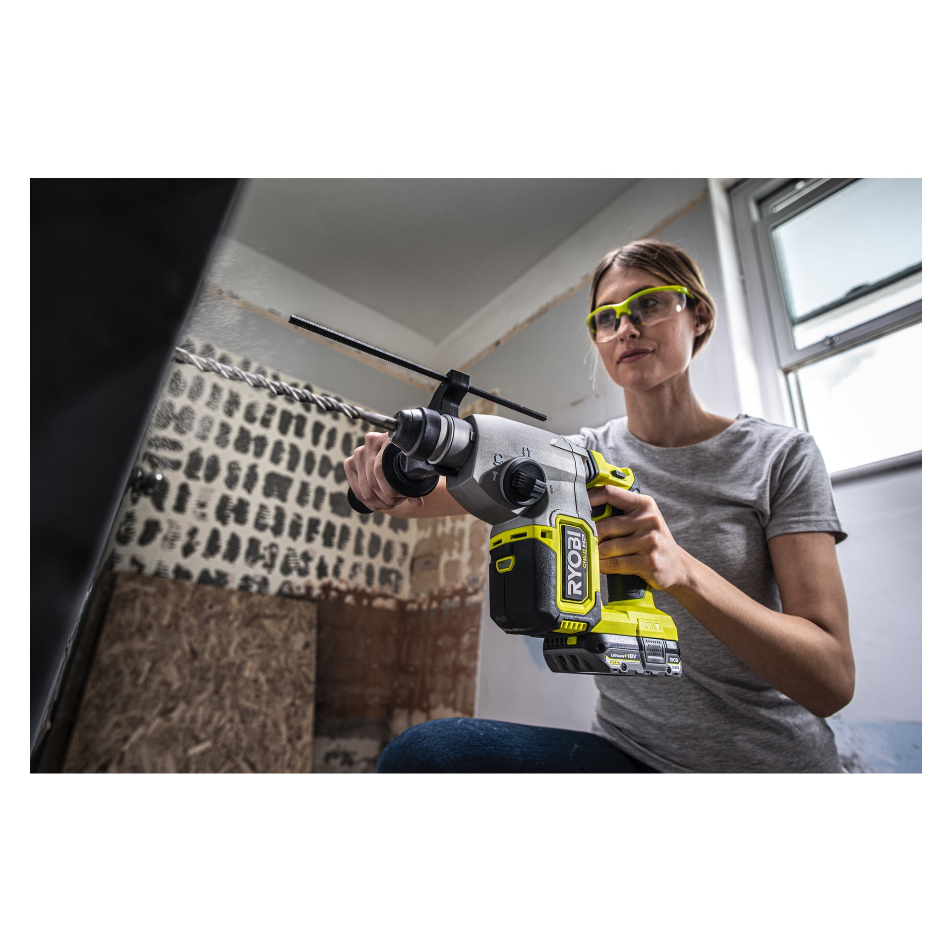 En person med skyddsglasögon använder en Ryobi One+ HP batteridriven hammarborr RSDS18X-0 i ett rum som renoveras, med ofärdiga väggar och ett fönster i bakgrunden.