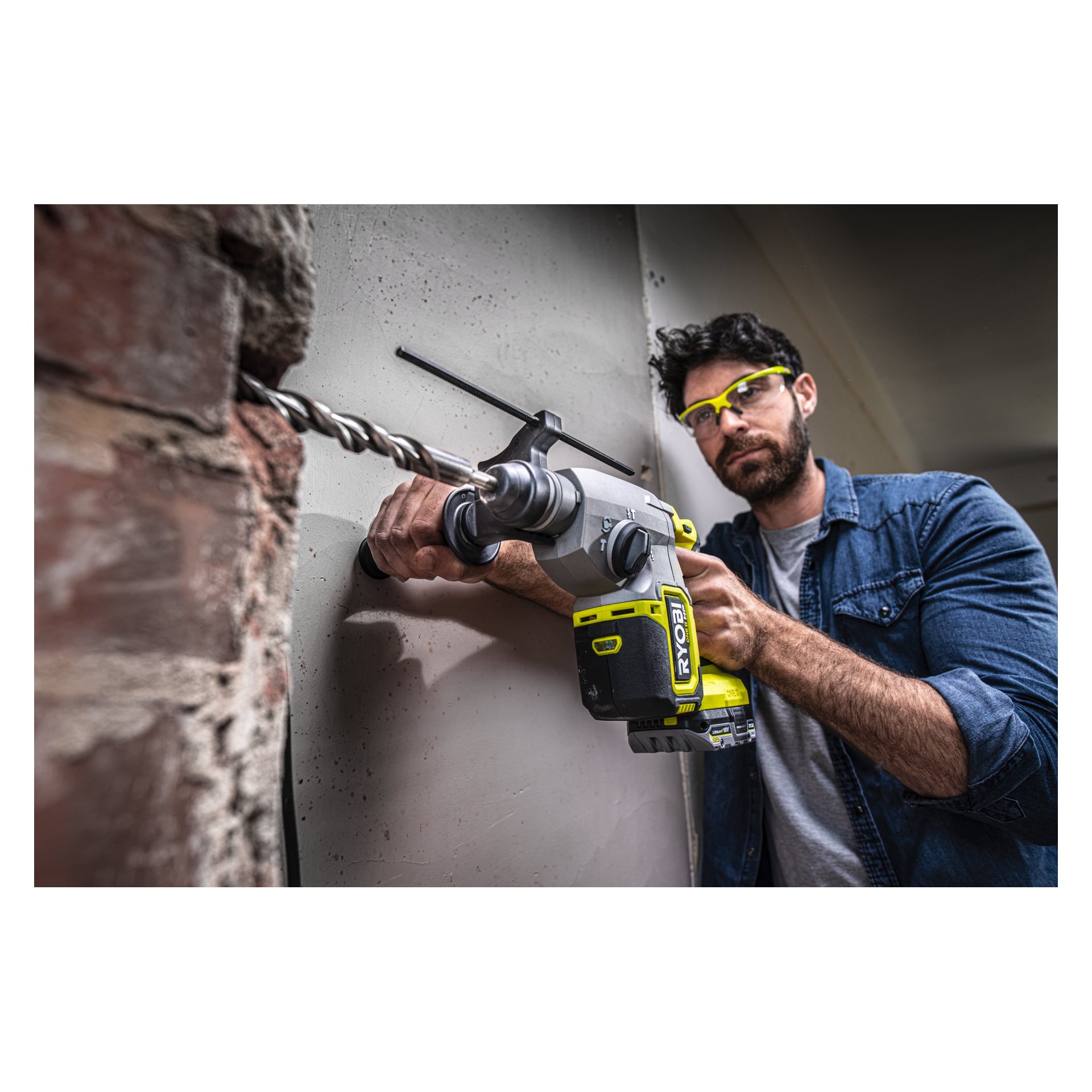 En man med skyddsglasögon borrar i en betongvägg med en Ryobi One+ HP batteridriven hammarborr RSDS18X-0. Han greppar verktyget stadigt med båda händerna och tittar intensivt på sitt arbete.